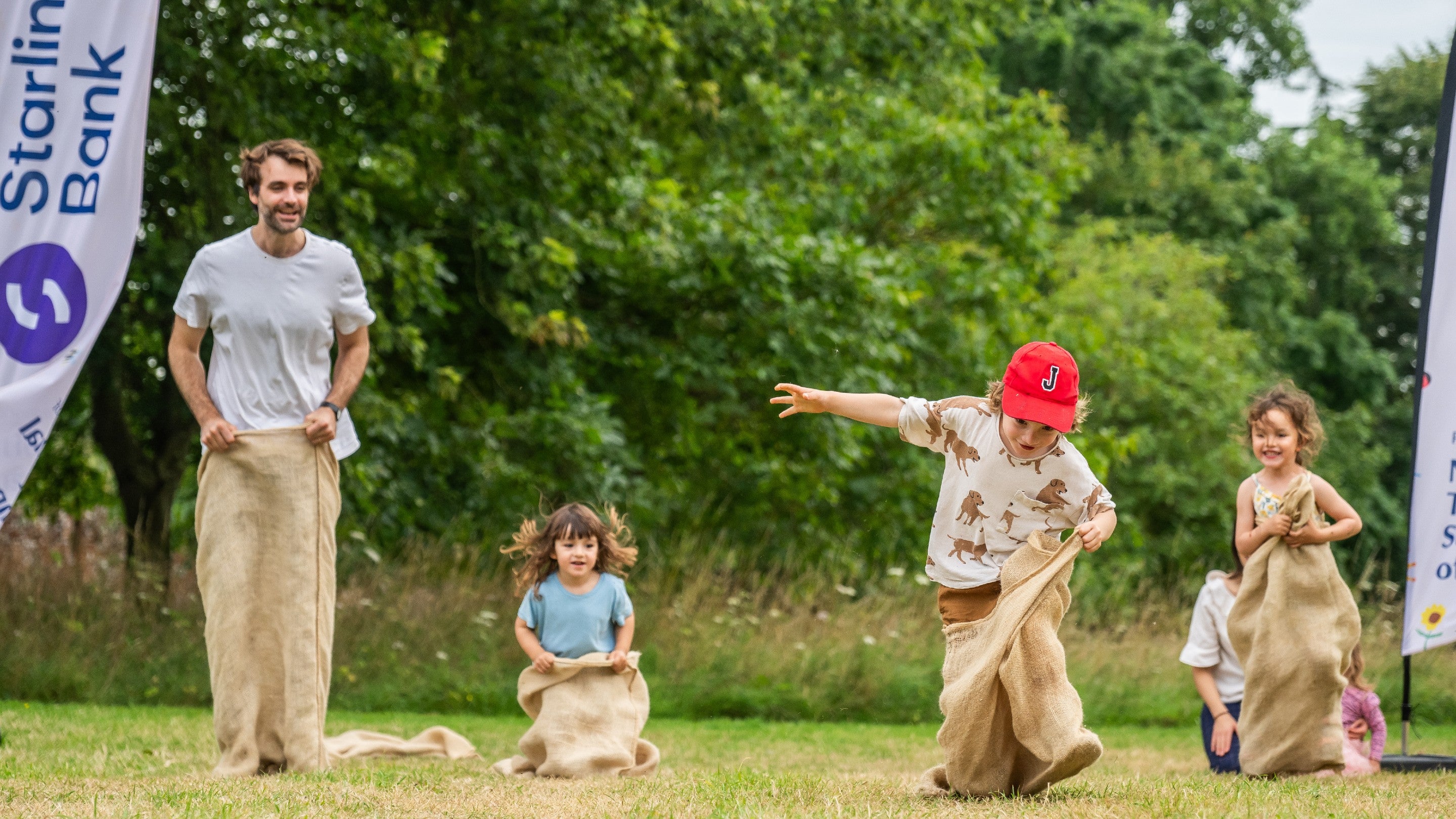 Dad and three children are jumping, taking part in a sack race outdoors. Smiles all round. There is a banner to the left-hand side reading 'Starling Bank'.