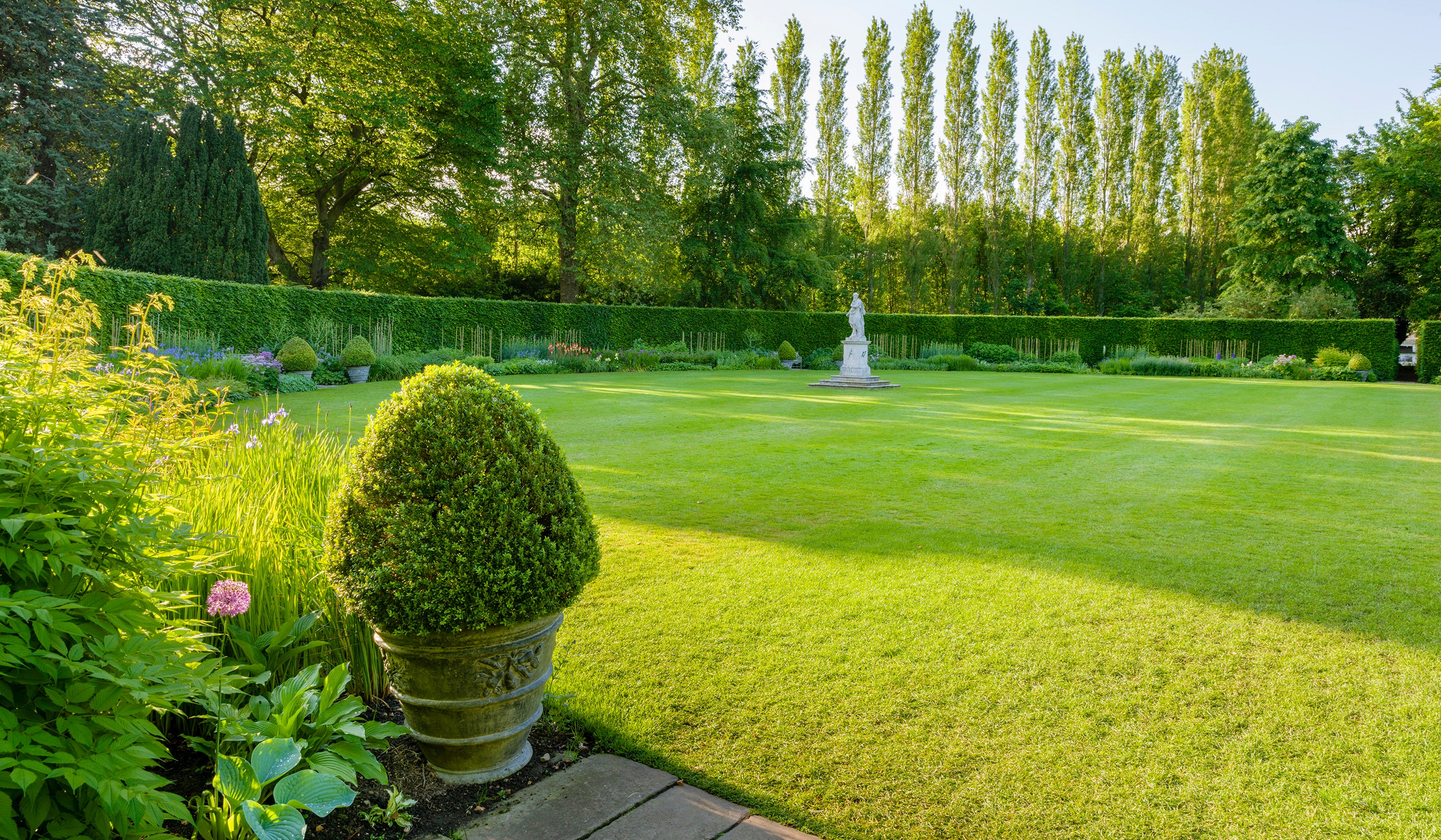 A photo showing a herbaceous border in early summer with flowers starting to bloom. There is a statue in the centre of a lawned area.