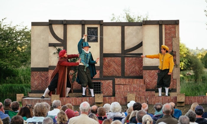 Three actors on a stage outside, dressed in red, blue and yellow costumes from left to right are performing a comical sword fighting scene as the crowd watches on.