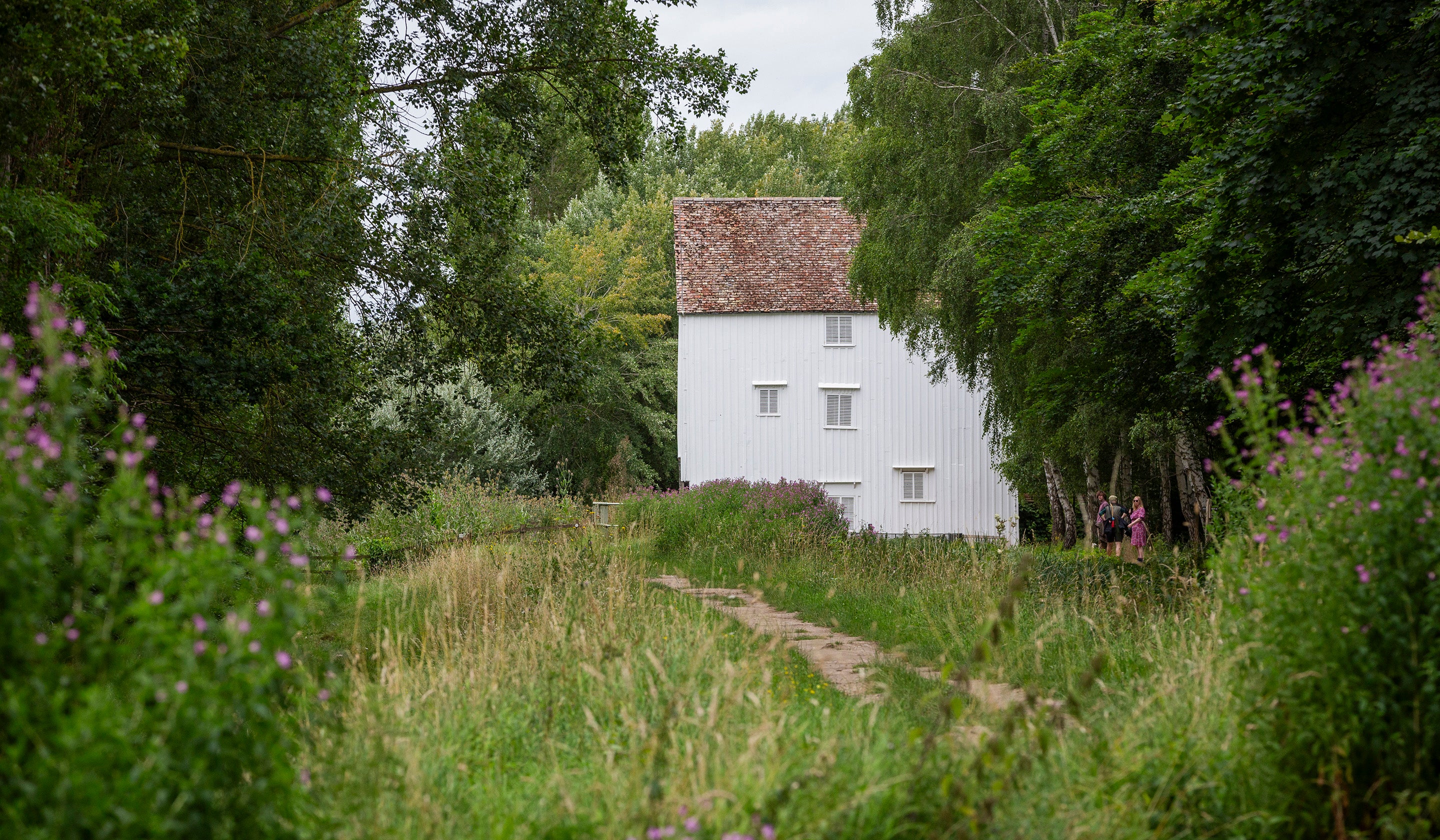 A view along a river towards a white clapboard mill building. It is summer and the foliage along the riverbank is lush and green