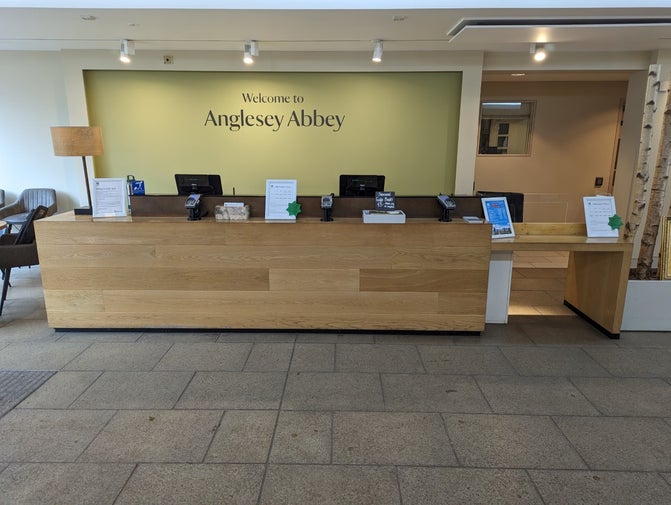 The welcome desk at Anglesey Abbey with till points and information stands. The desk is in front of a green wall that says 'welcome to Anglesey Abbey'.
