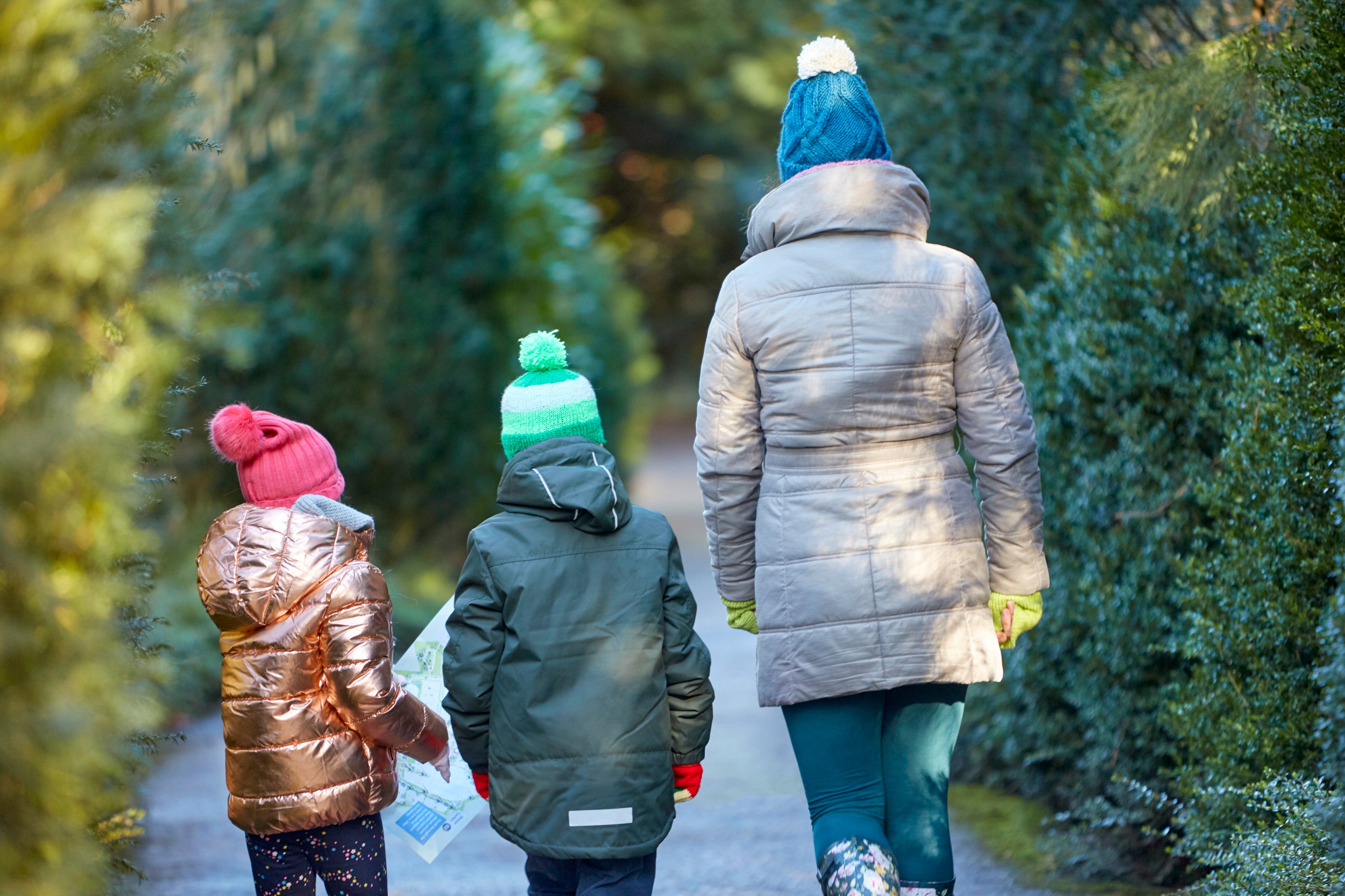 A mum and two children walking through the gardens at Anglesey Abbey with a map in hand, backs to the camera with woolly hats on