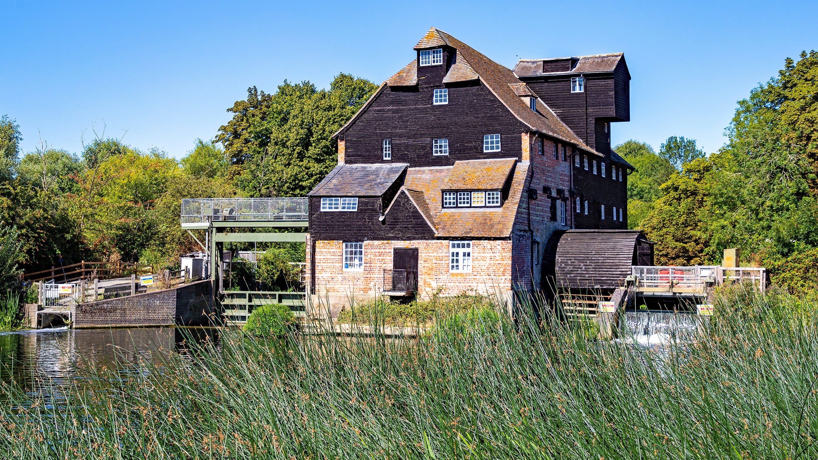 An historic riverside watermill, built of red brick and black boards. It is summer in the photo, the sky is blue and the riverbank is green with vegetation