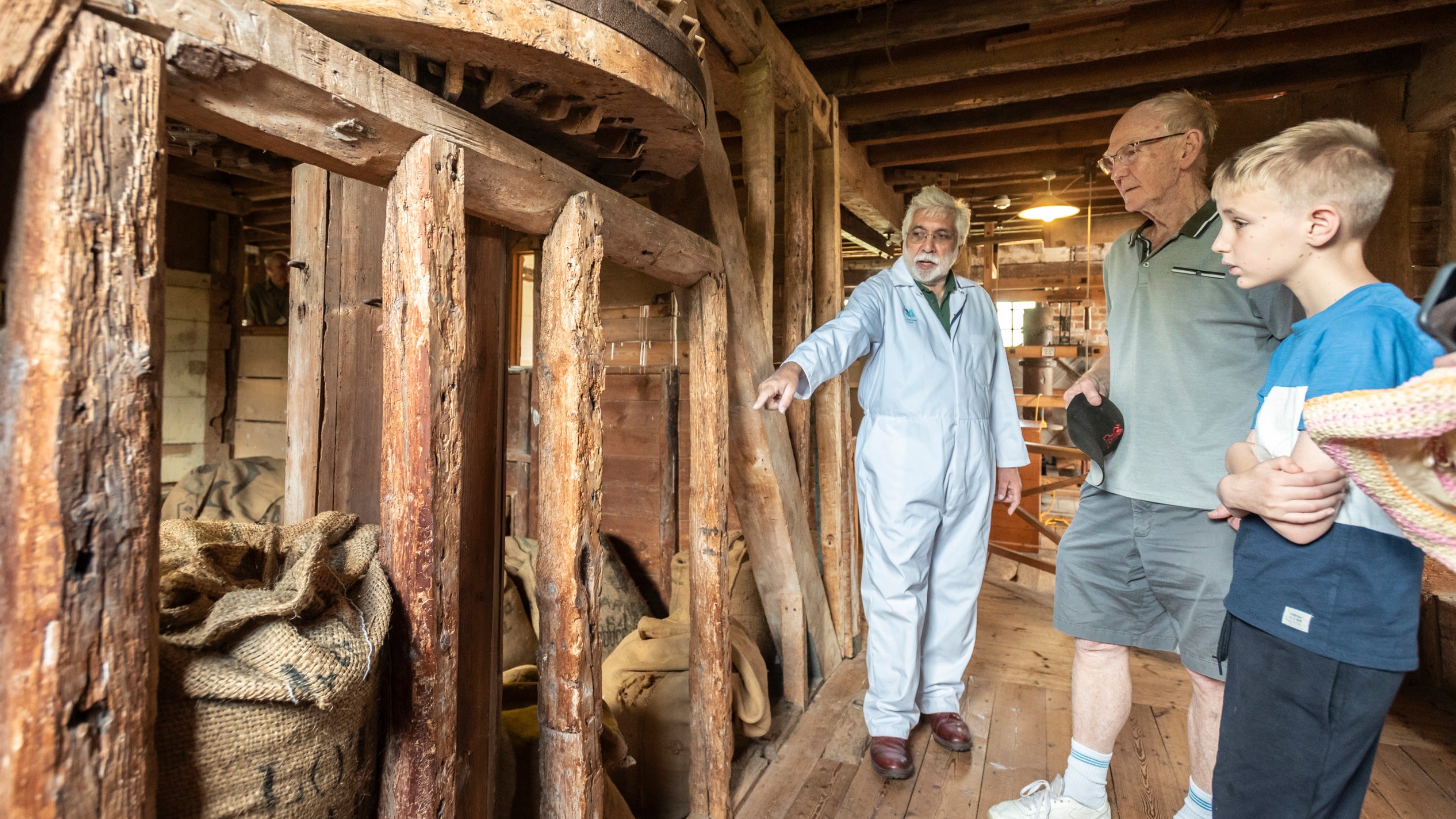 Wooden mechanisms inside the mill including a turning wheel and bags of grain with visitors and a volunteer looking.