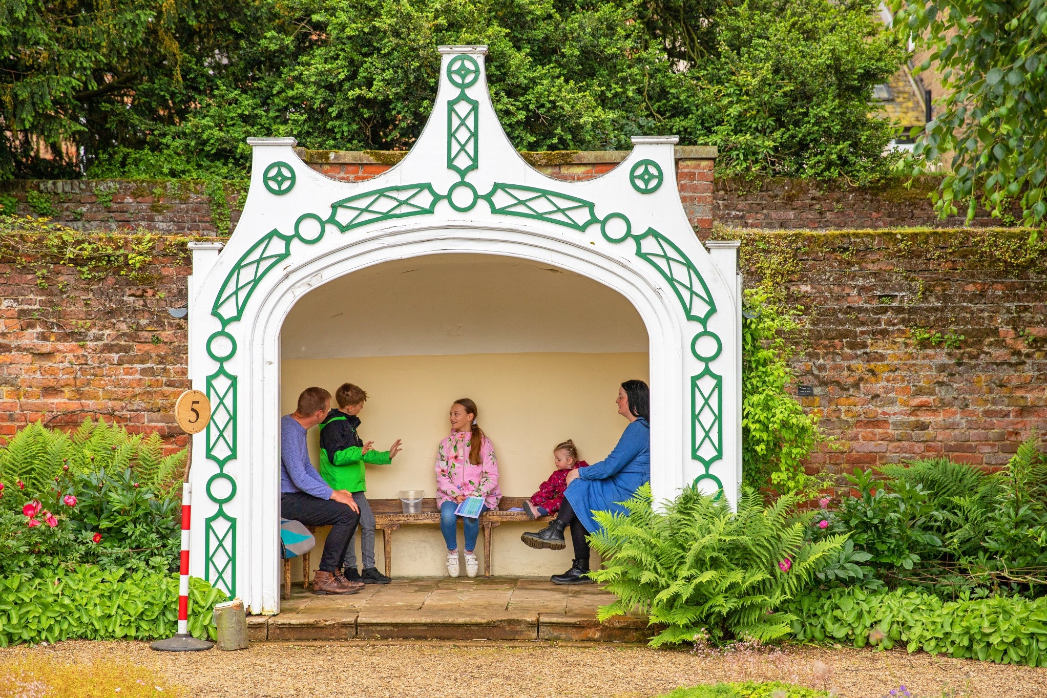 A family of five sit in the stone summer house at Peckover to shelter from the rain. They are laughing with each other.