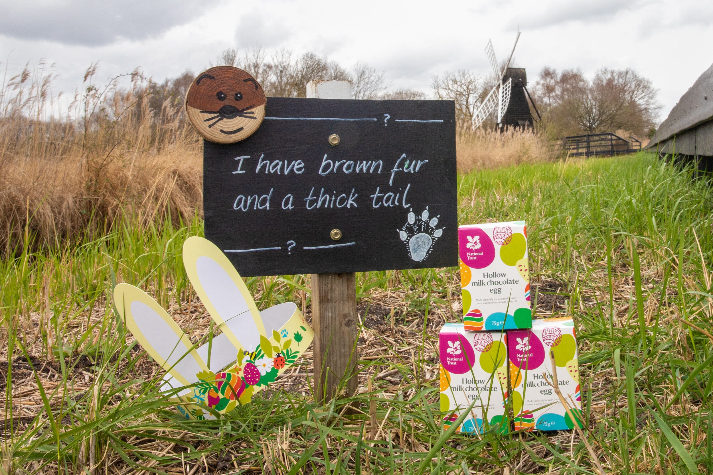 Easter eggs, bunny ears beside boardwalk and a and sign post for 'Easter Trail at Wicken Fen' with greenery and windpump in the distance