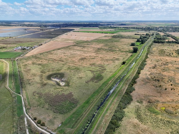 An aerial view across green fields, with scattered trees and a waterway running through the middle