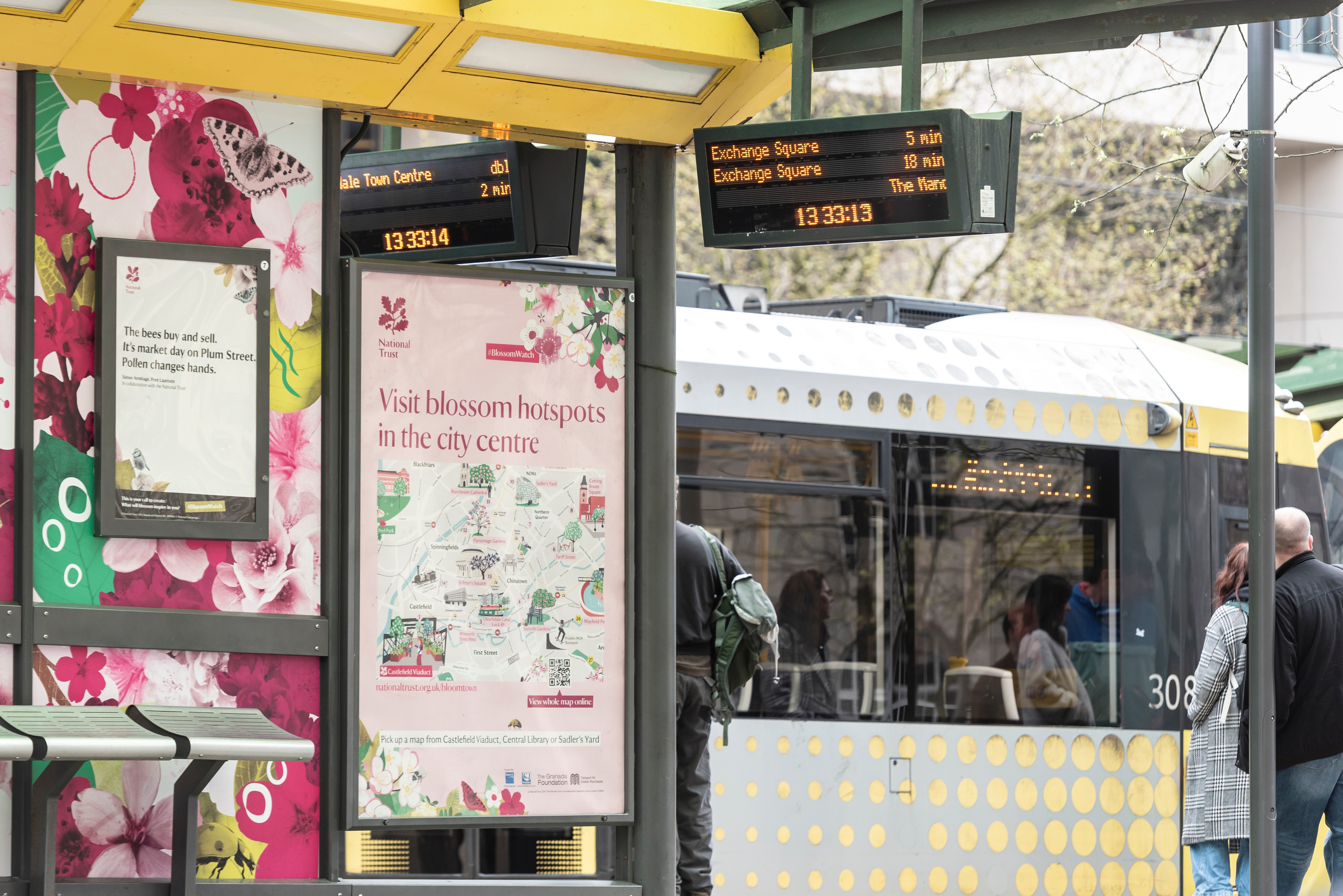 The Bloomtown Map is displayed at a tram stop at St Peter's square in Manchester. Behind is a tram which is decorated with pink blossom and leaves.