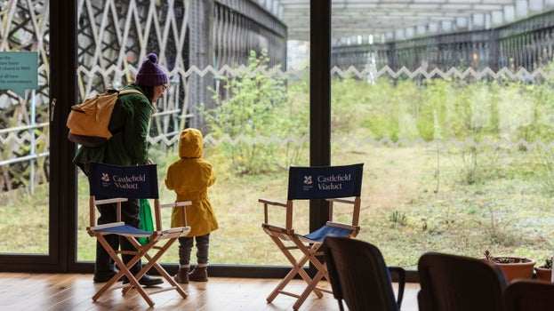 Two people look out of a glass window to a Victorian viaduct overgrown with green plants.