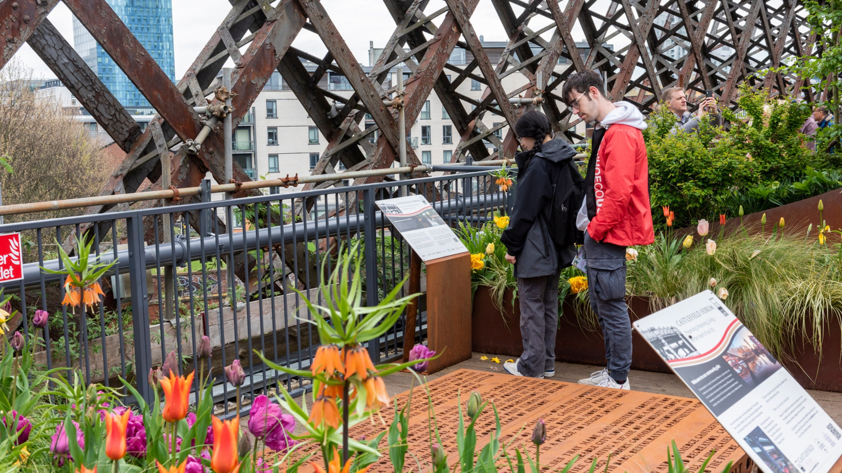 Visitors on the Bloomtown Trail in spring at Castlefield Viaduct, Greater Manchester, read a sign about the trail which pinpoints blossoming trees in and around Manchester, with colourful spring flowers in the foreground