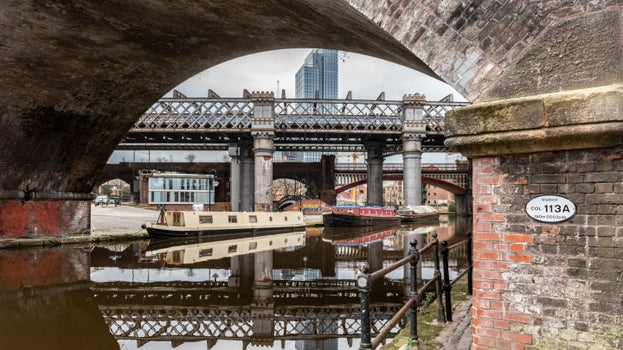 A view through the arch of a bridge over the canal, with canal boats and their reflections in the water, and Castlefield Viaduct visible in the background with other city features.