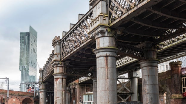 A photograph taken from the ground beneath Castlefield Viaduct looking up at the steel structure, with a skyscraper visible in the background.