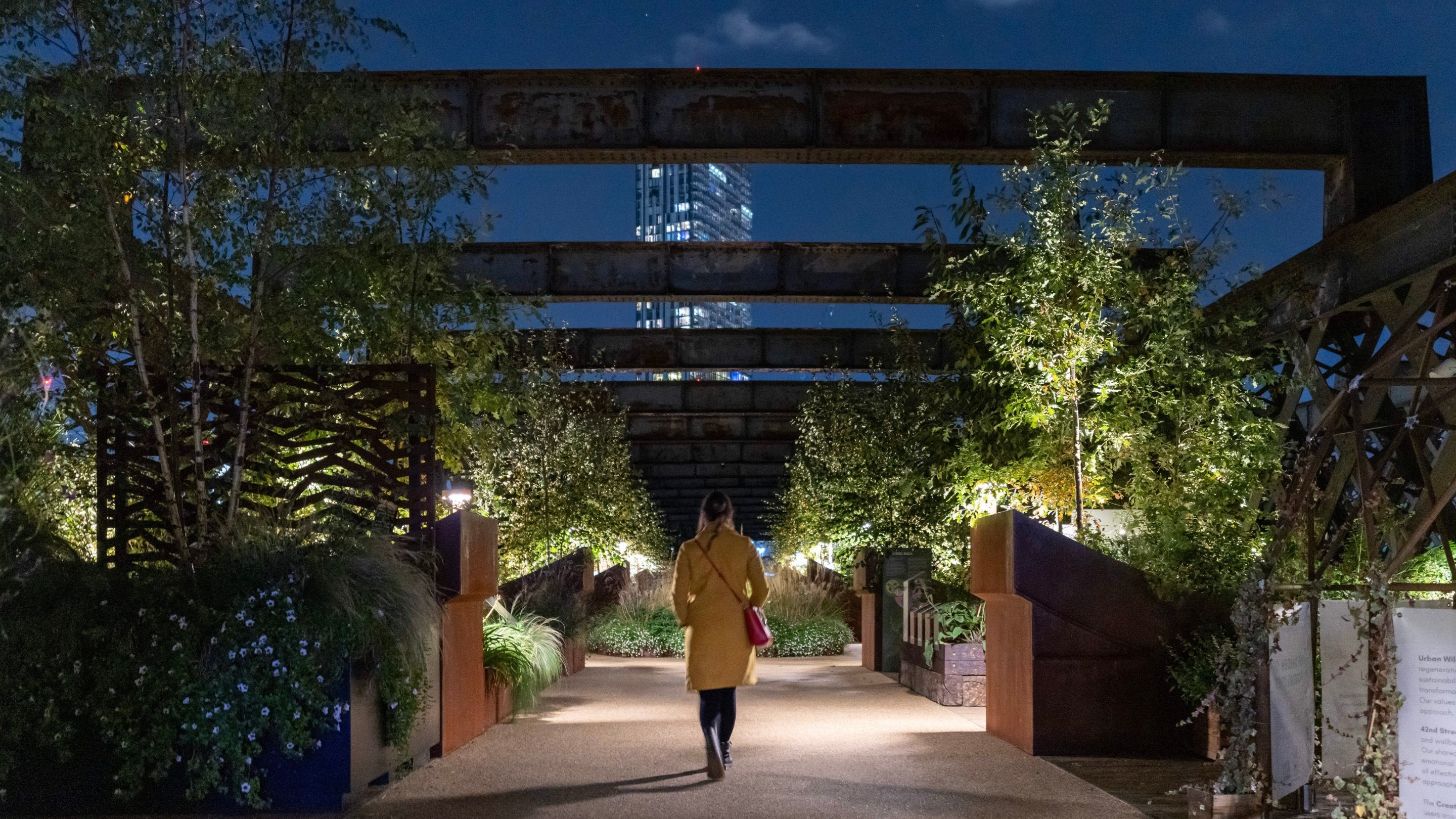 A woman in a yellow coat walking down Castlefield Viaduct in Manchester at night surrounded by lanterns in the gardens.