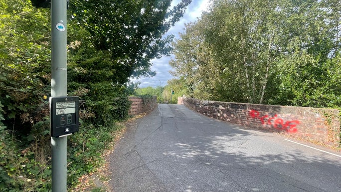 An image of a pedestrian crossing of a narrowed road across the canal with a button on the left hand side.