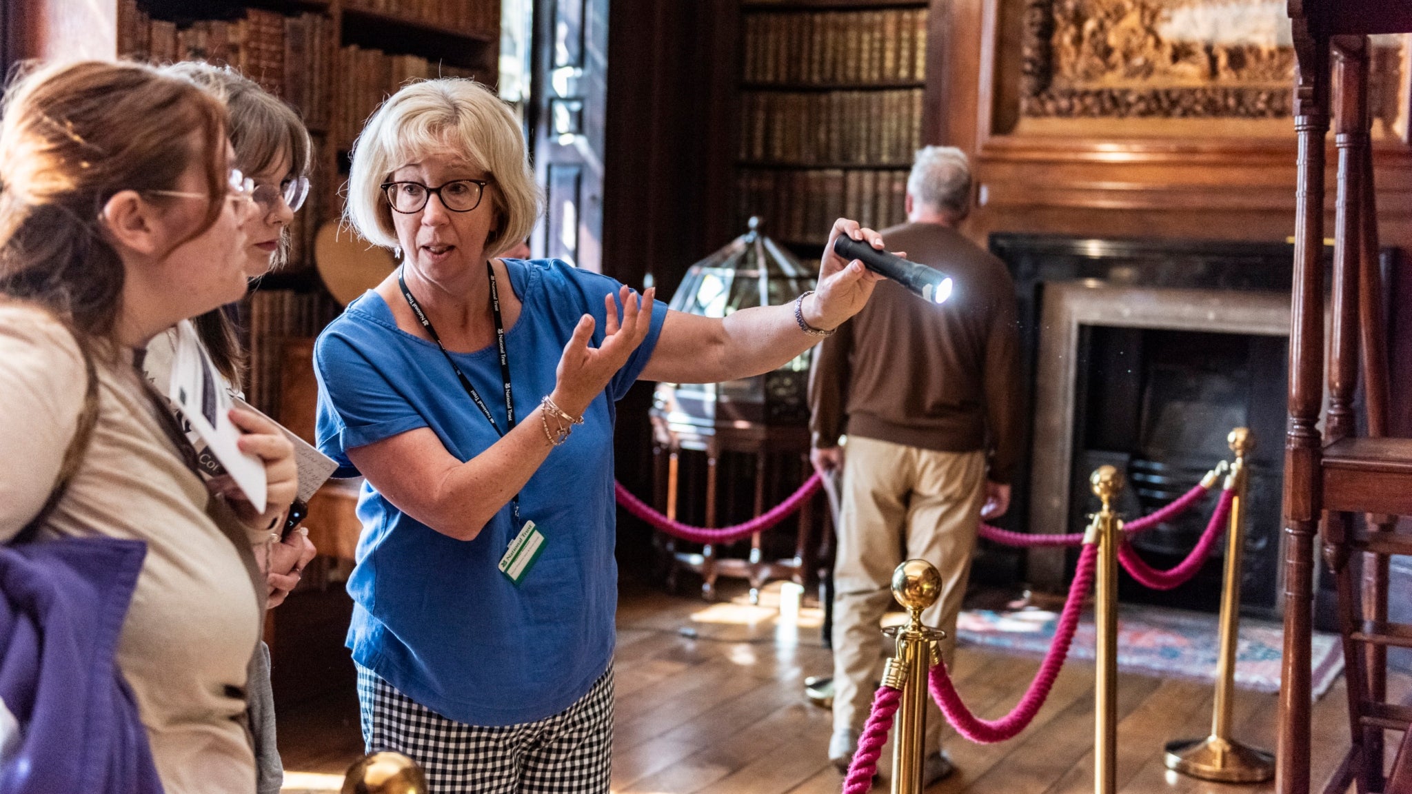 A woman wearing a volunteer lanyard is seen talking to visitors, pointing to an item on the shelf, in the Libary at Dunham Massey, Cheshire