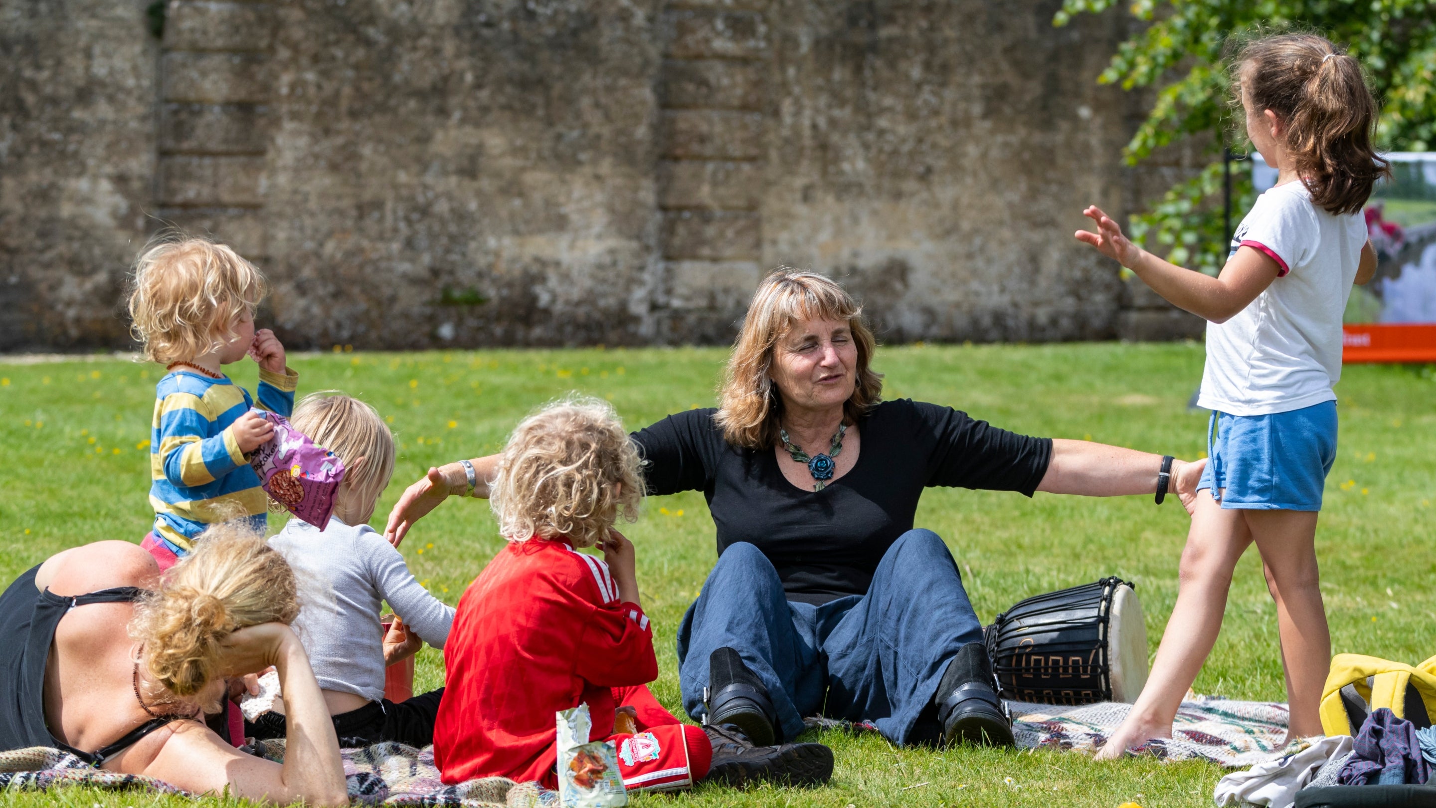 A lady and children sitting on the grass in a park, engaged in storytelling and enjoying a sunny day together.
