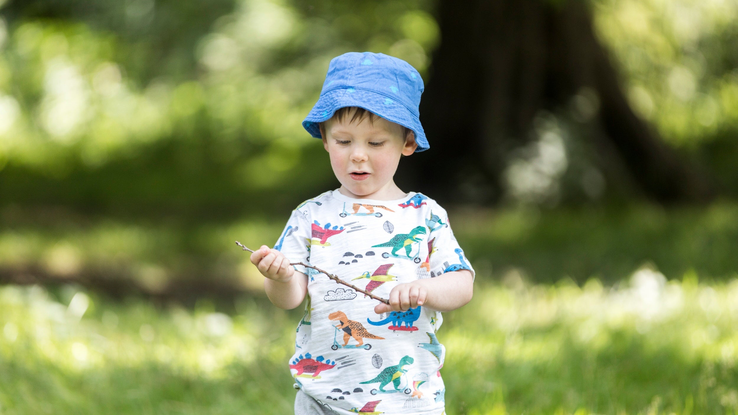 A small child in a blue sunhat playing with a twig, surrounded by green trees