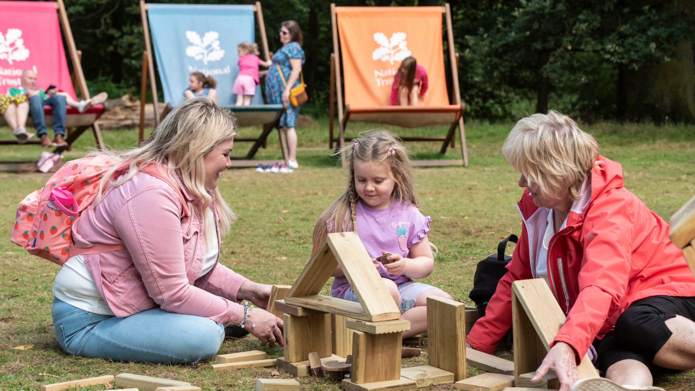 A family sat on the grass, playing with some wooden blocks. Giant deckchairs can be seen on the background and people sat on them.