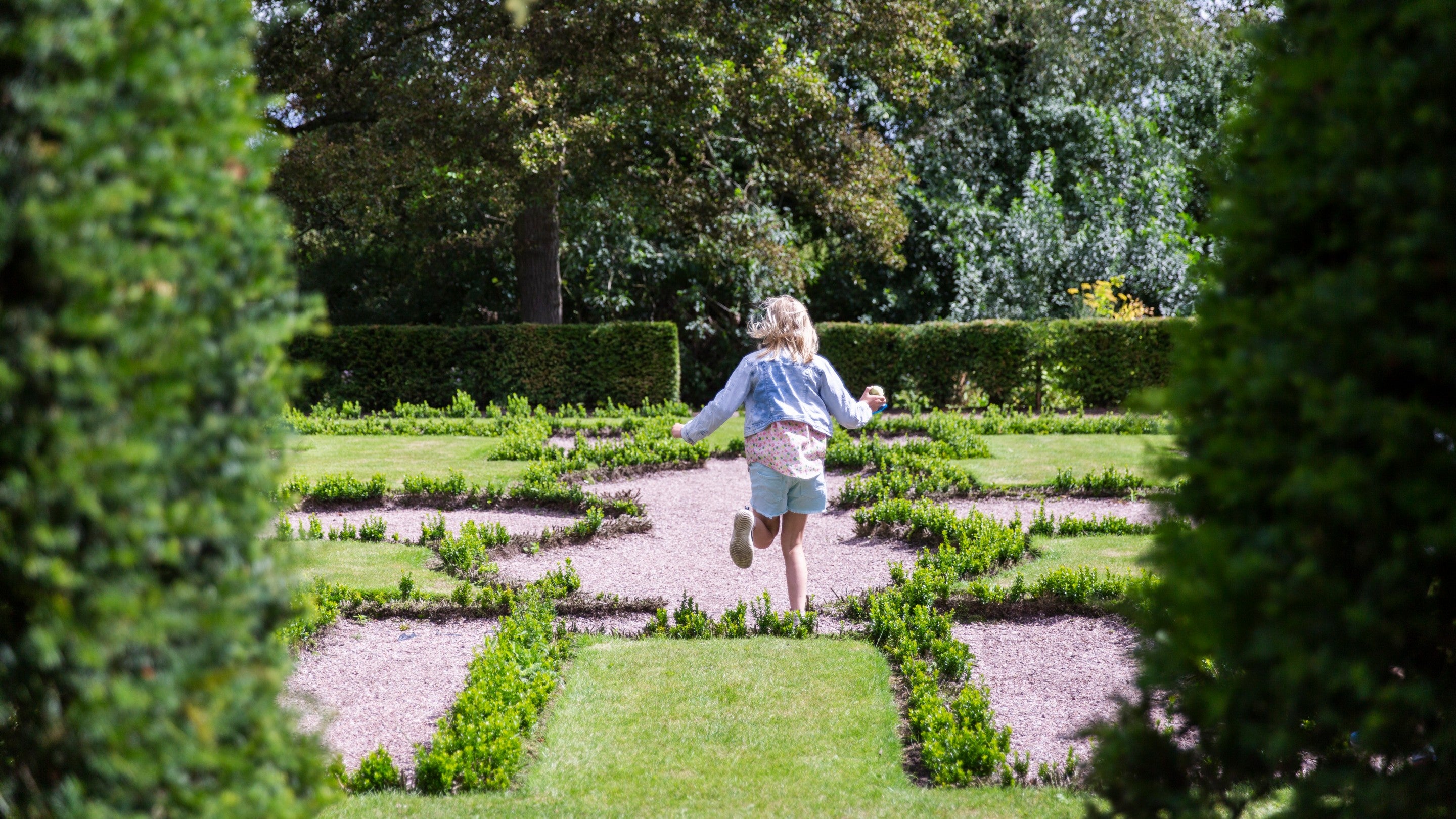 A girl in a sunhat runs into a symmetrical garden at Little Moreton Hall, Cheshire