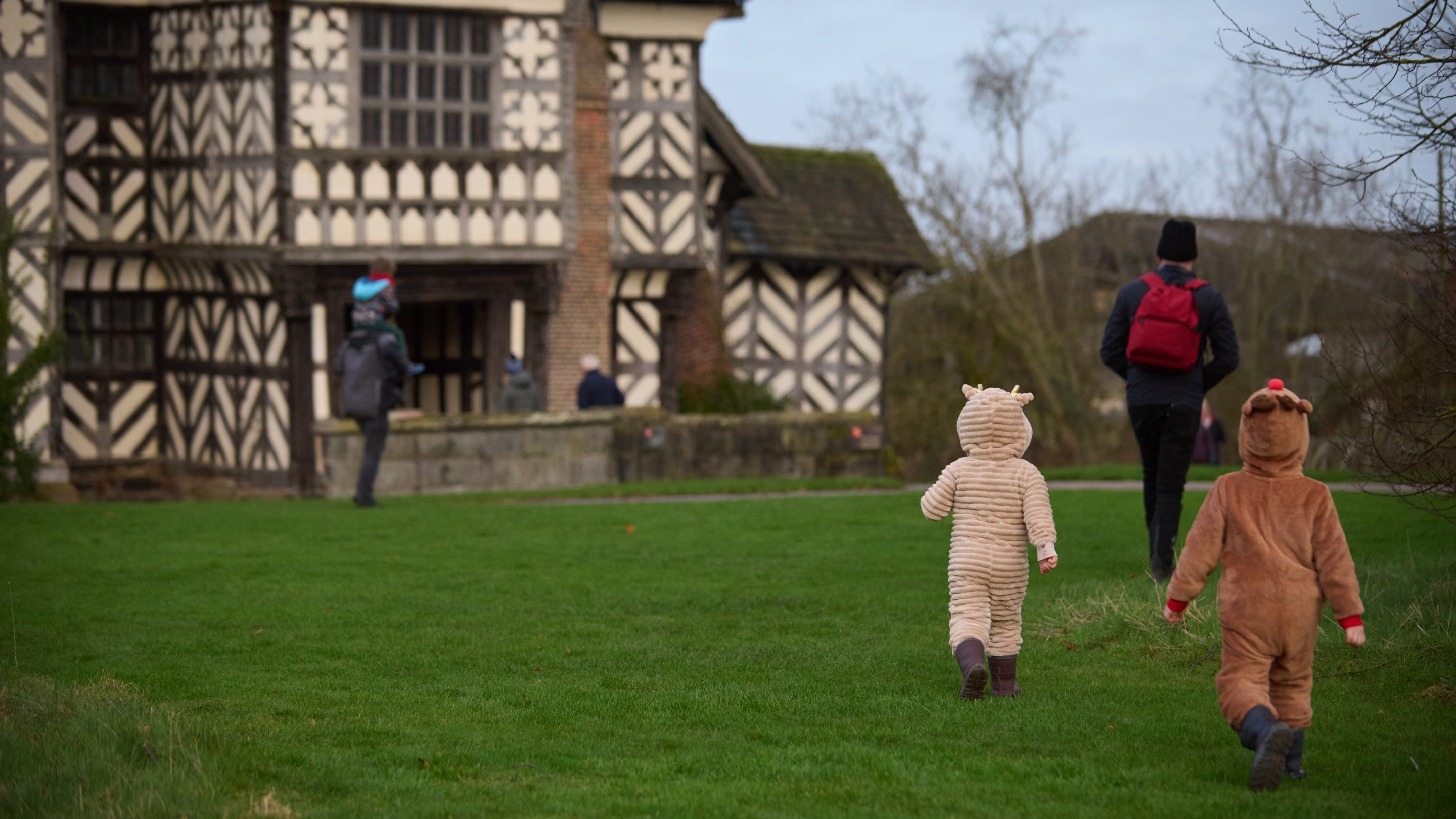 Two children dressed in reindeer onesies walk over grass towards a black and white Tudor house.