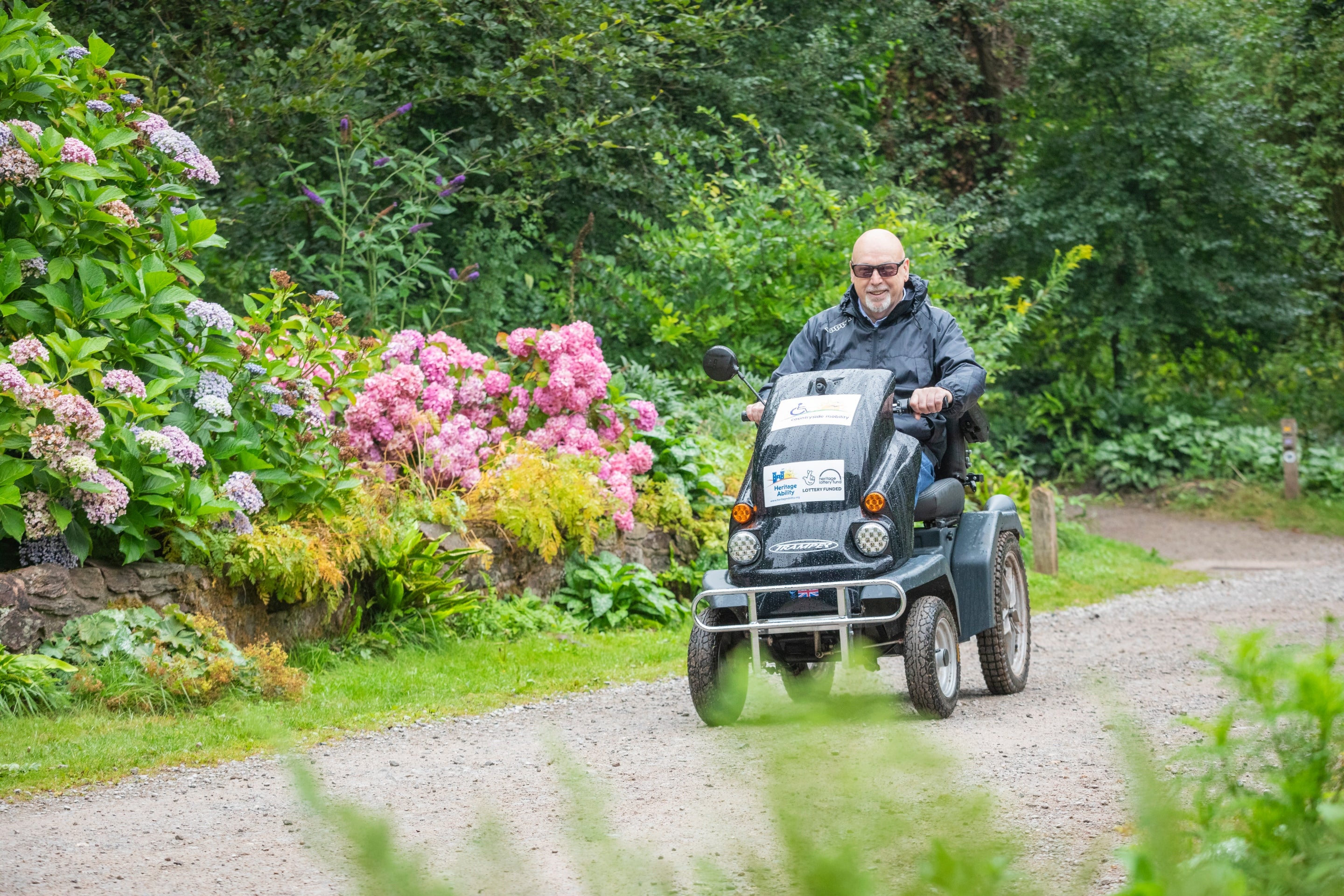 A visitor explores gardens in a tramper, a four wheeled mobility vehicle designed for uneven terrain. Pink and purple flowers and green foliage frame the image.