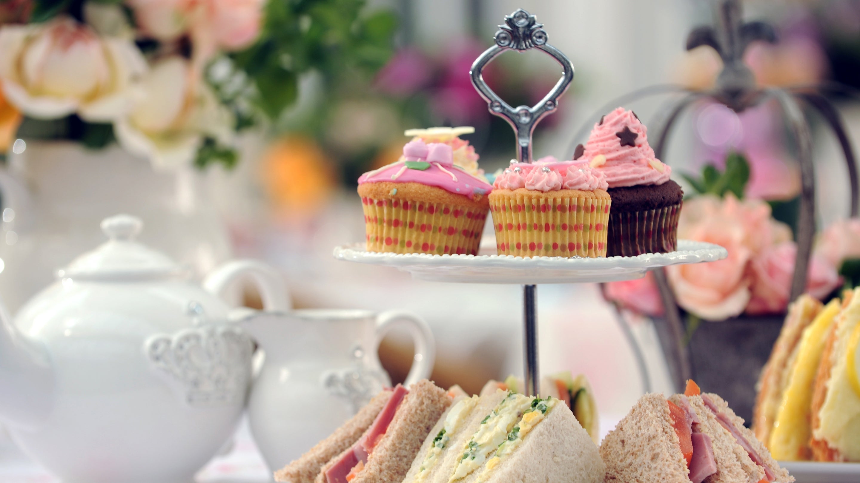 A tiered afternoon tea with iced cakes and sandwiches, set against colourful flowers.