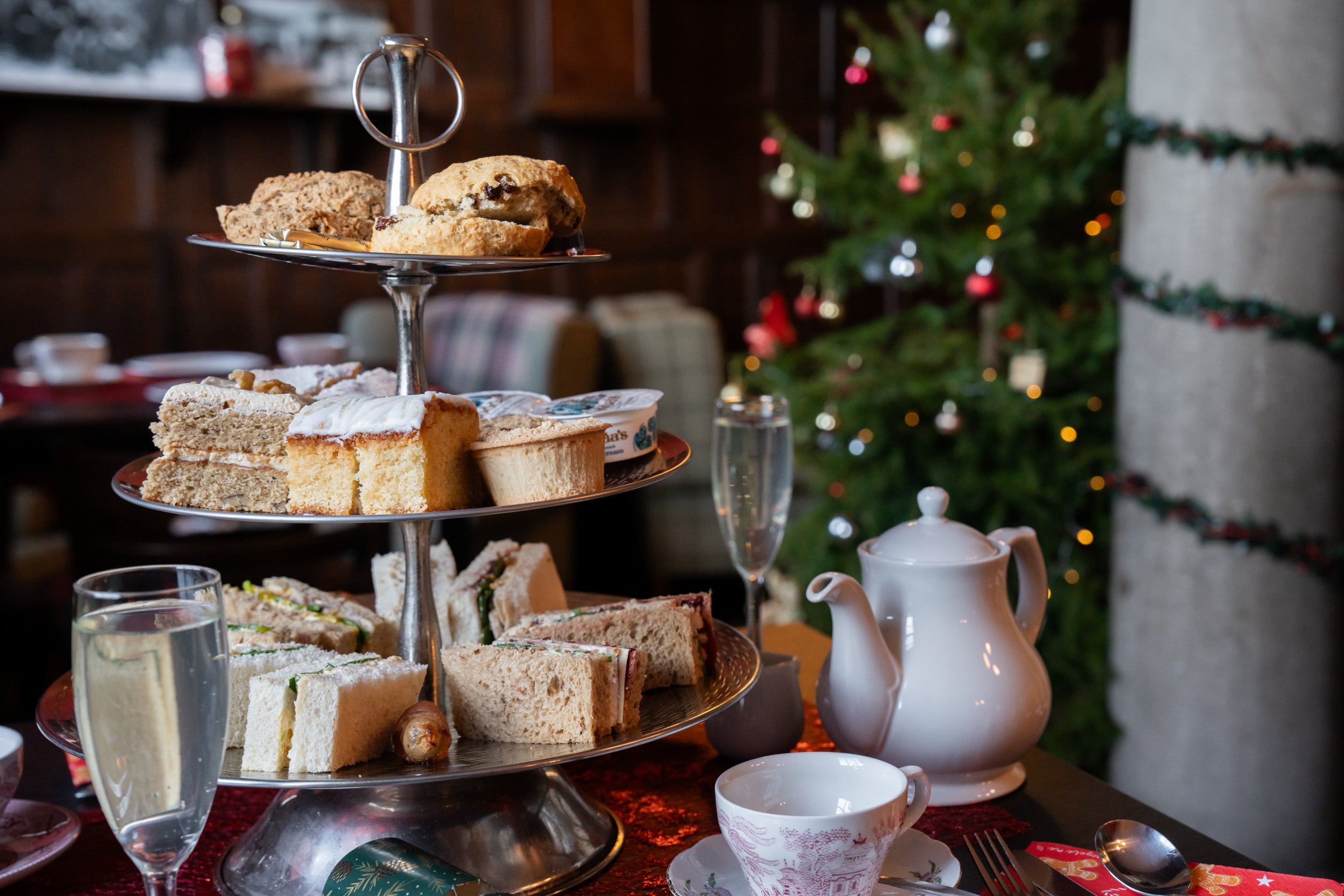 A tiered afternoon tea stand brimming with cakes, sandwiches and scones sits on a table with glasses of elderflower bubbly, a teapot and a cup of tea. There is a decorated Christmas tree in the background as well as a pillar wrapped in a festive garland.