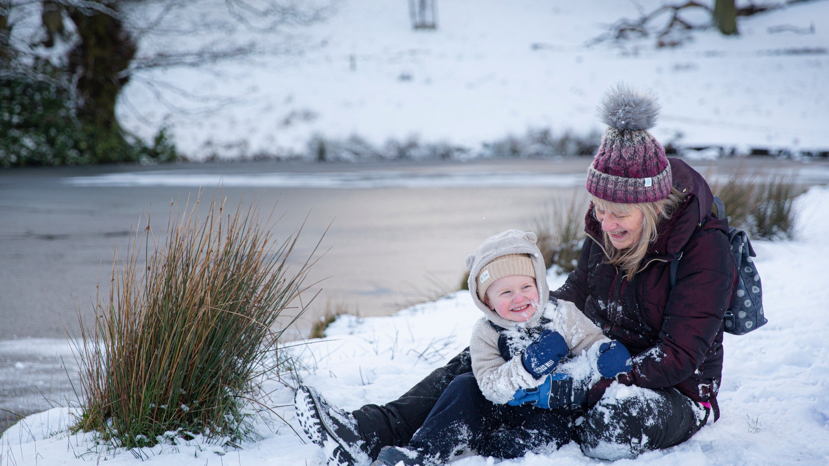 A woman and toddler lay playing in the snow next to a frozen pond.