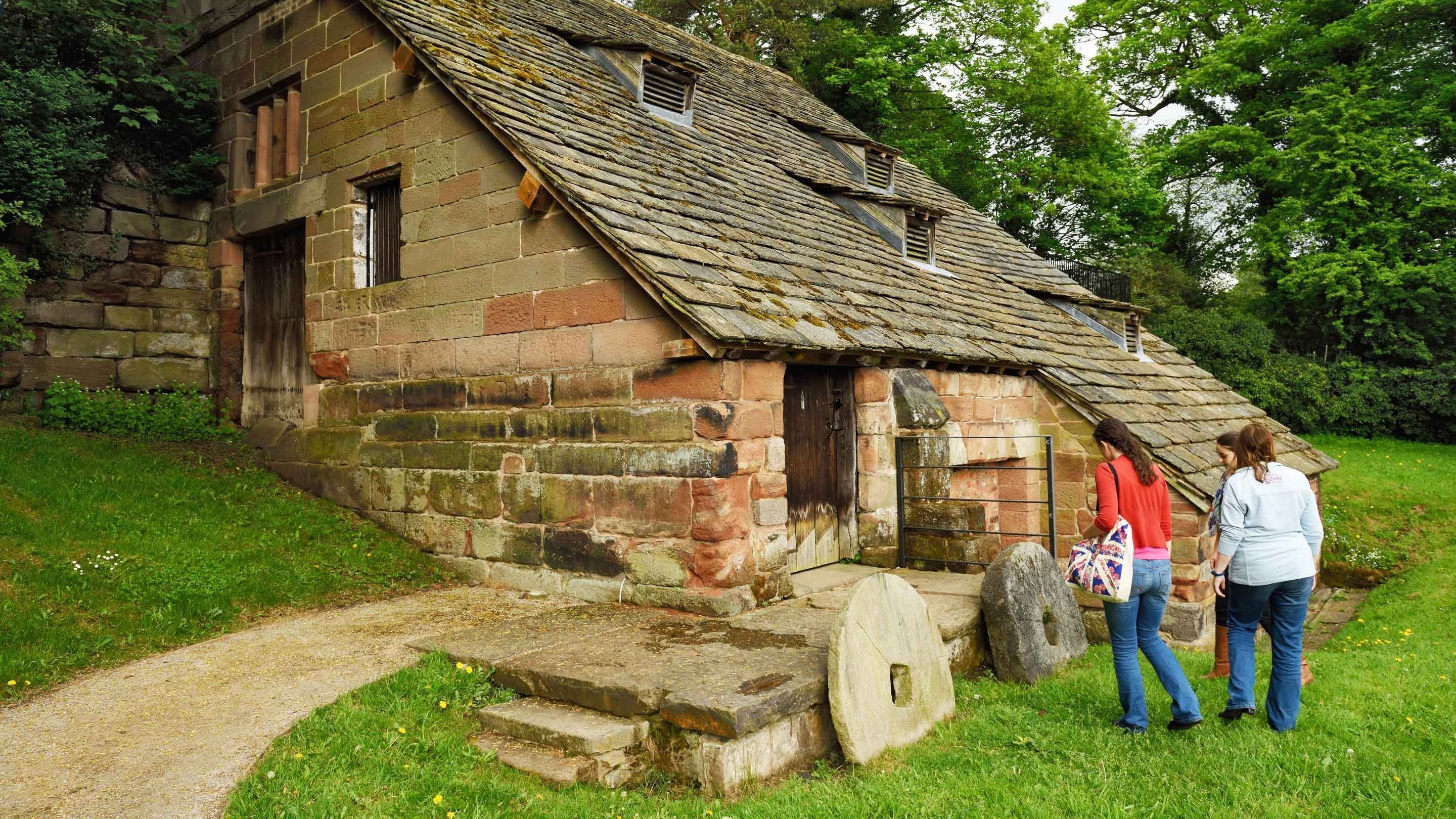 Three visitors are looking at two large circular millstones which are leaning against the mill building at Nether Alderley Mill, Cheshire. The building's roof slopes steeply down almost to the ground.