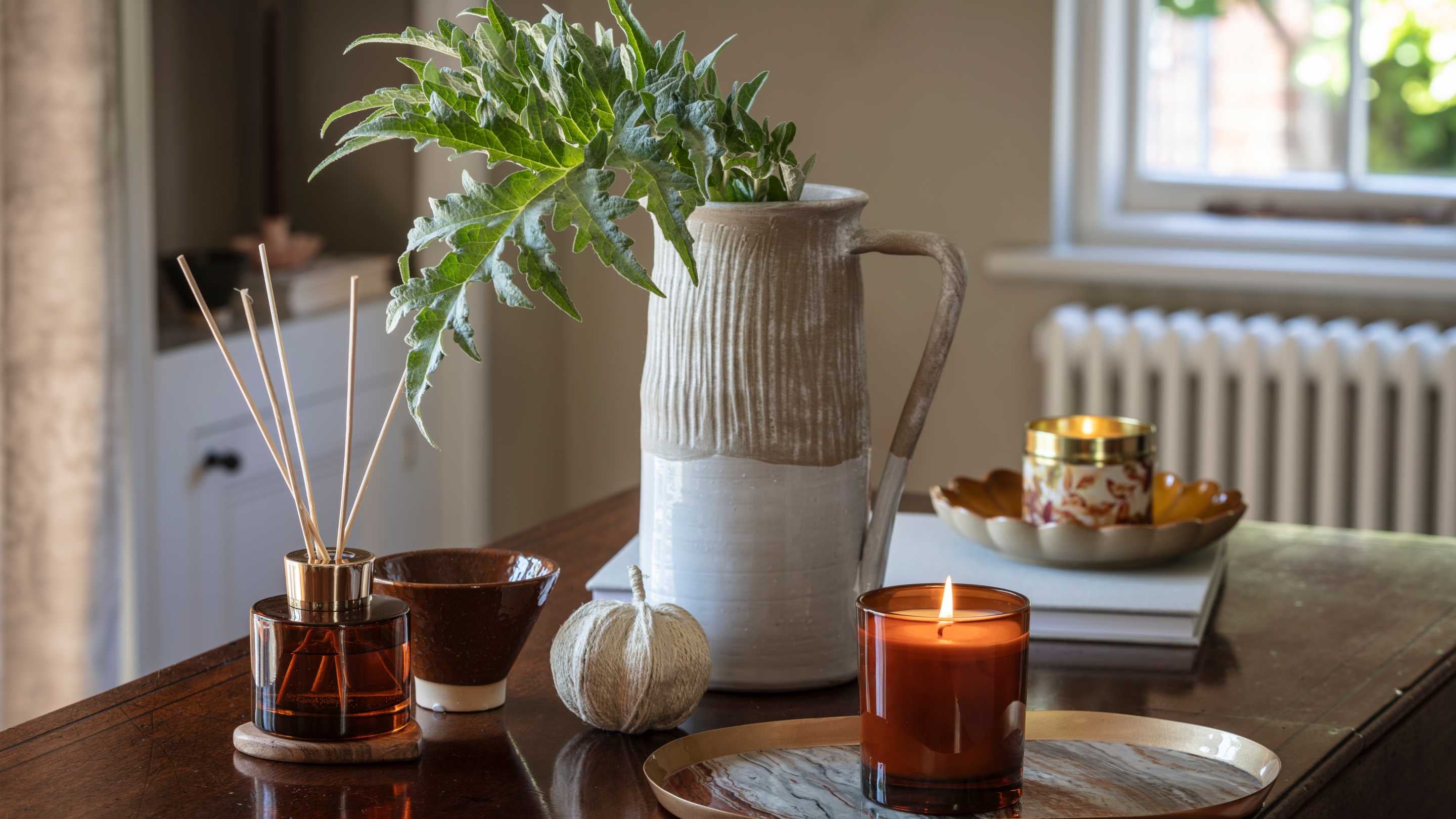A table with a jug with leaves displayed, candles and a reed difuser