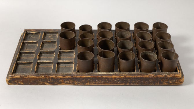 A wooden tray with squares carved out of it, in the holes sit round metal cups