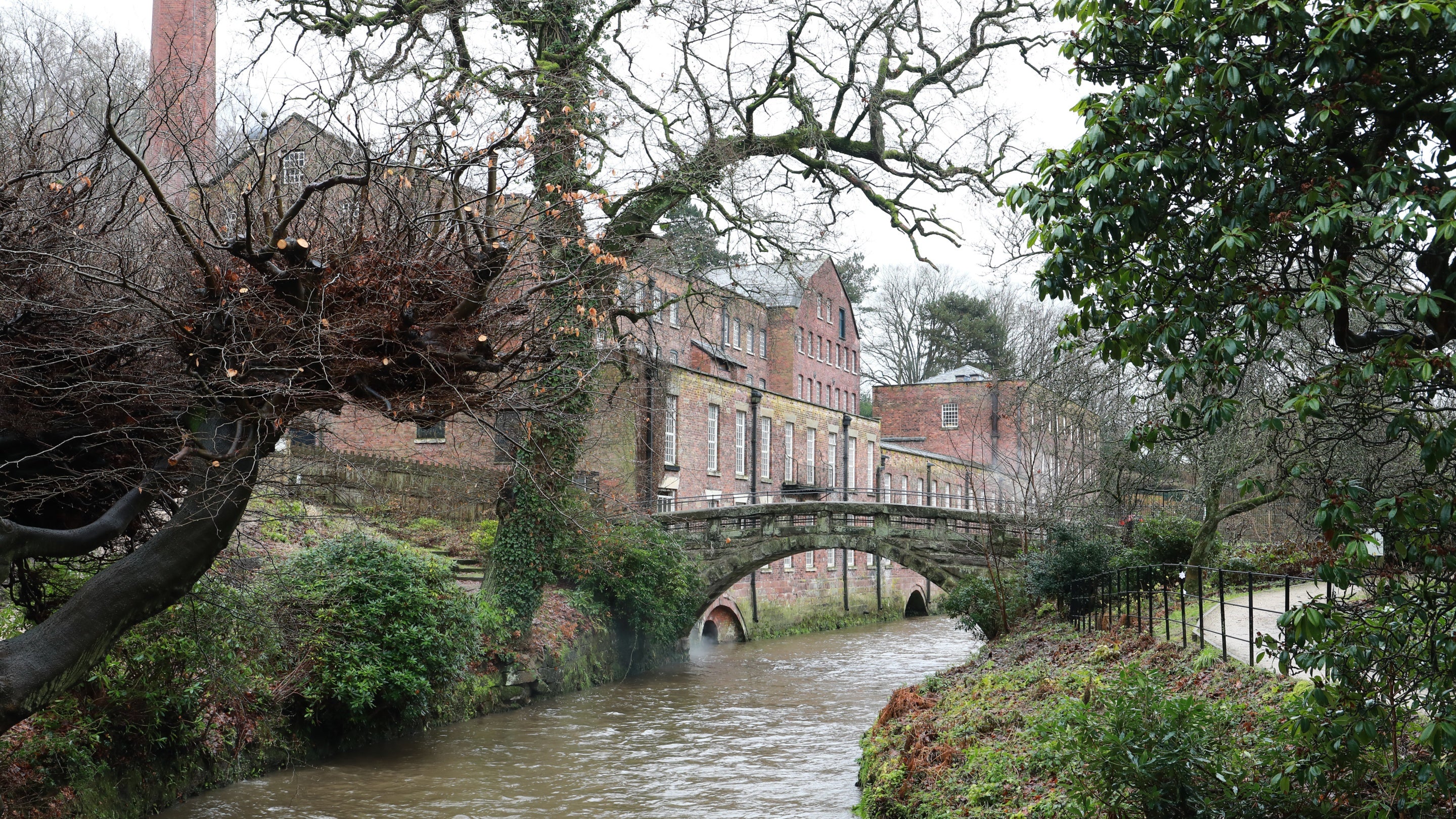 Long red brick mill building with stone bridge and brown river water below