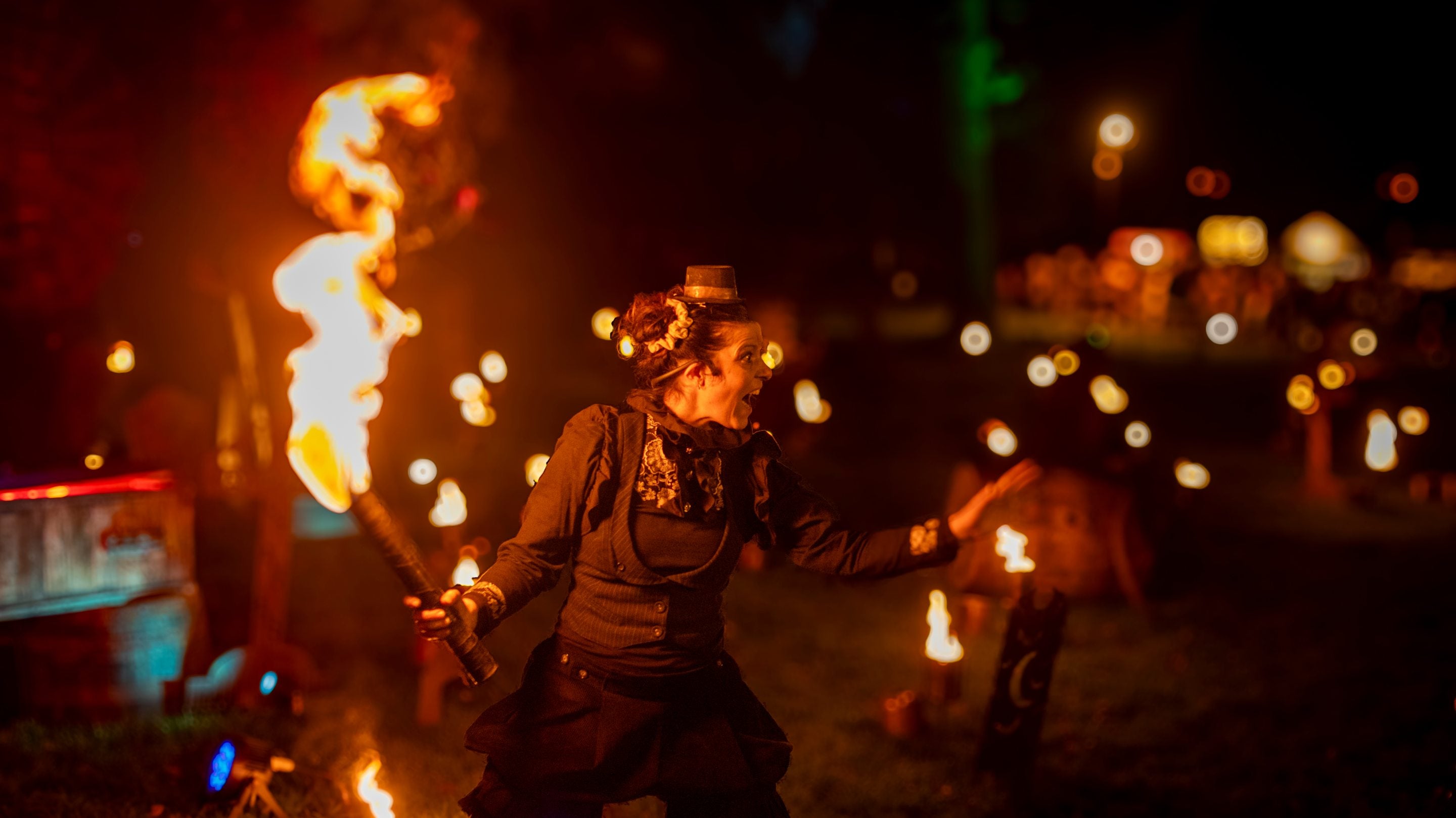 Night time scene with figure dressed in black with fire lanterns in the background. The perormer is holding a stick on fire