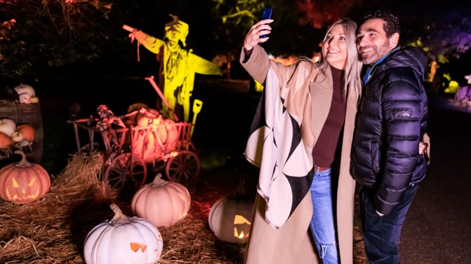 A man and a women embracing as they take a selfie. In the background a scarecrow looms and pumpkins glowing on the floor.