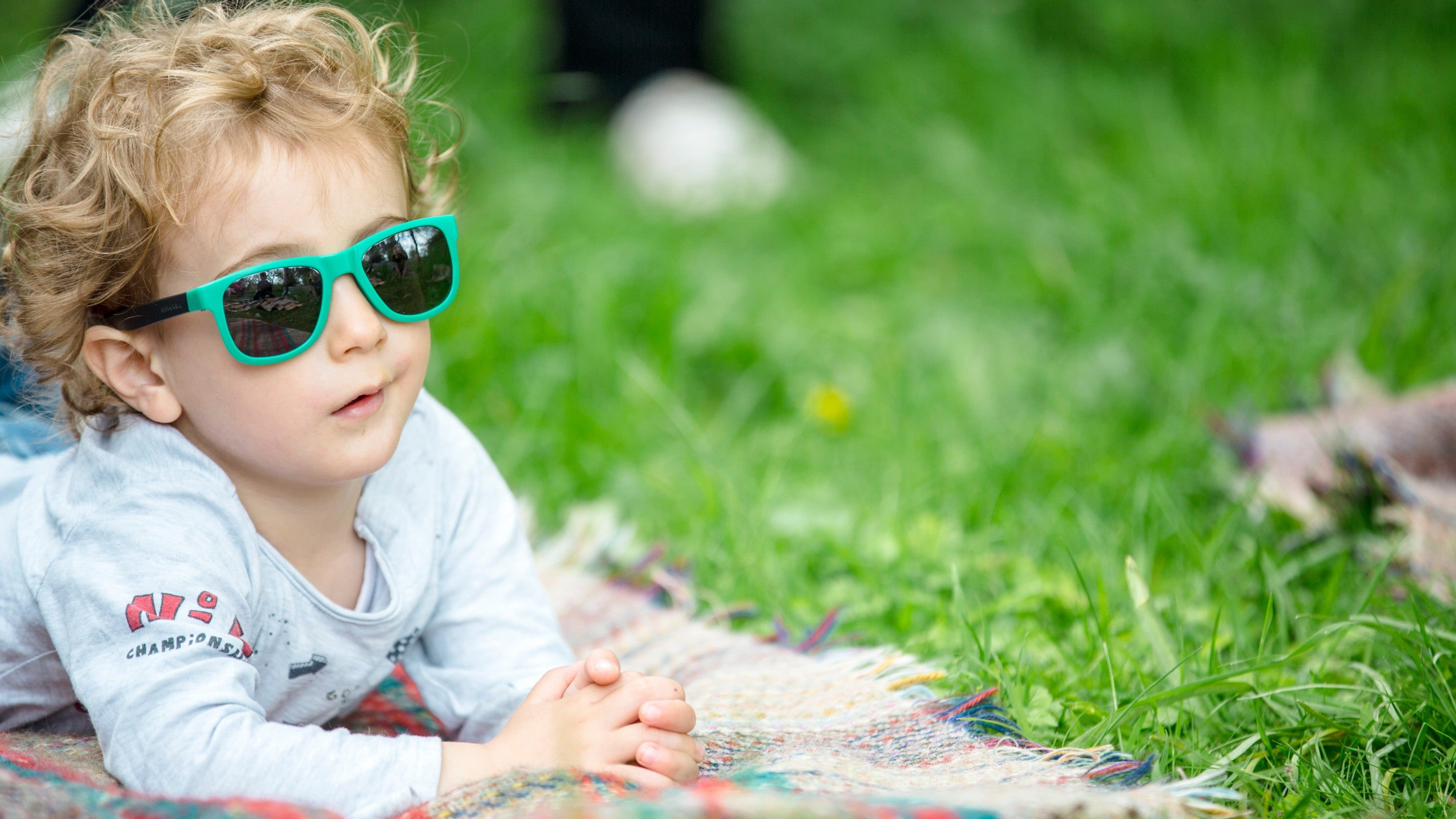 A child with curly blonde hair wearing green sunglasses lying on a picnic blanket.
