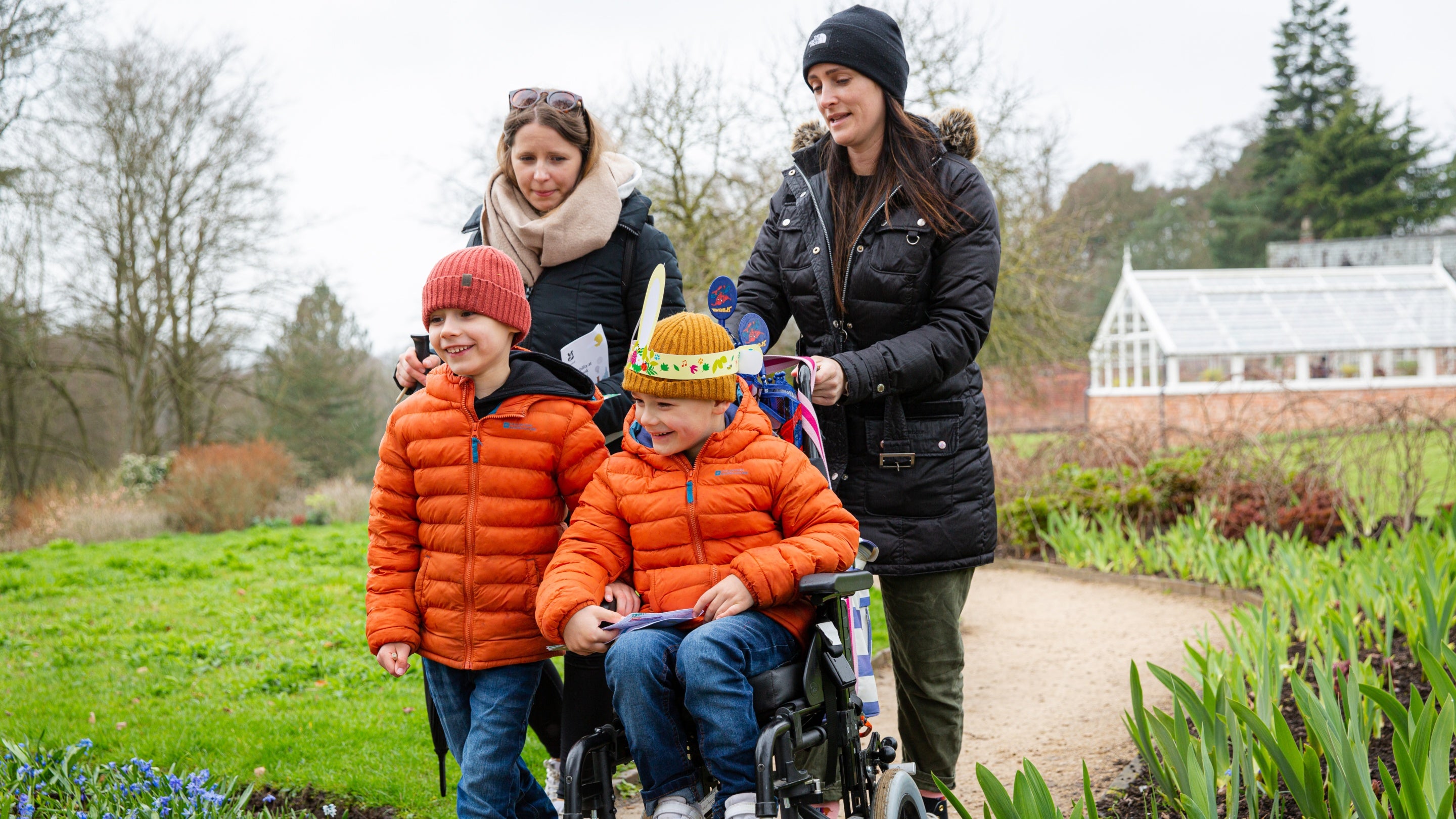 Two children, one in a wheelchair with two women walking through garde with a glasshouse in the background