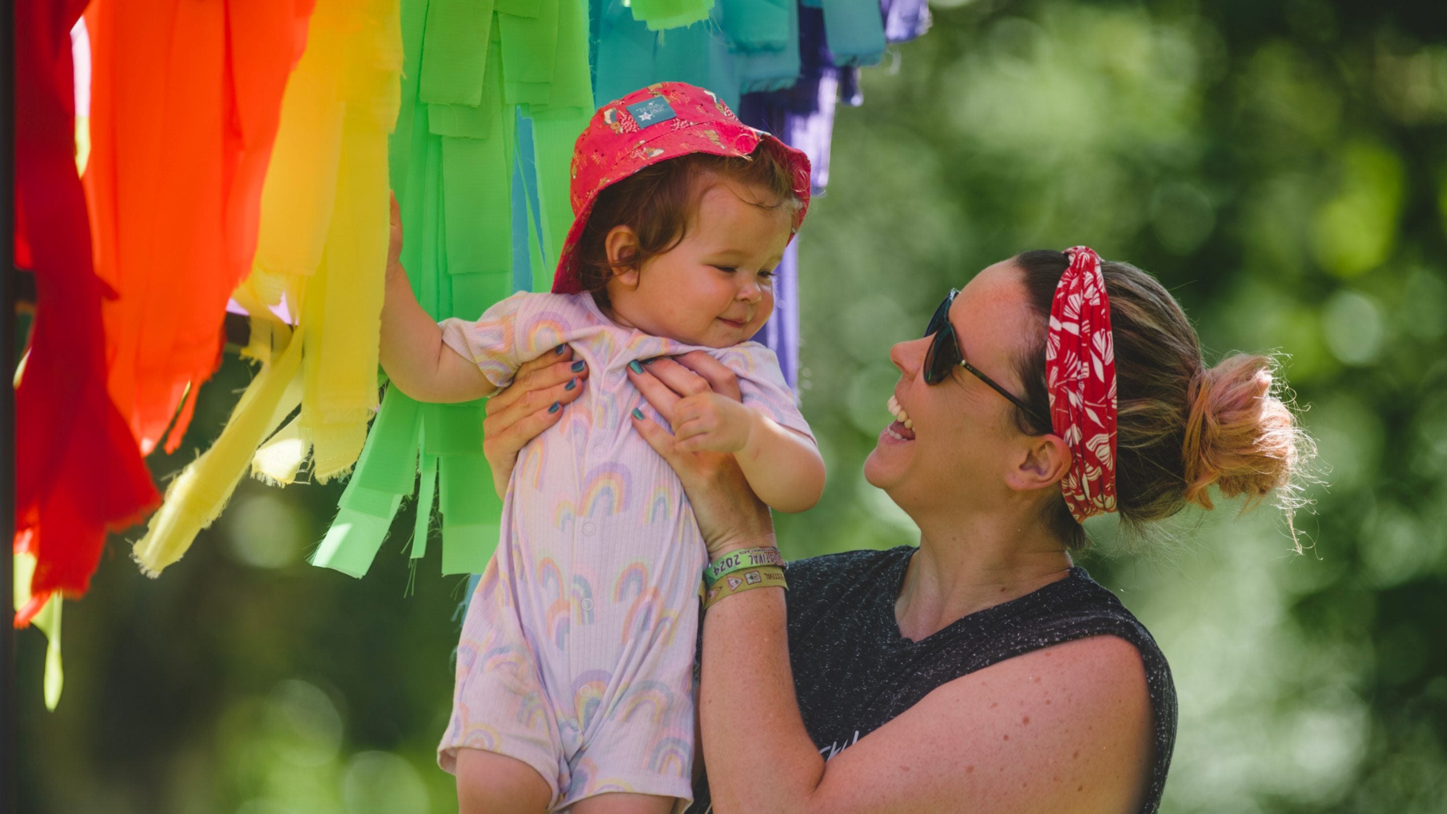 A woman holding a babay wearing a sun hat with rainbow of ribbons in the background