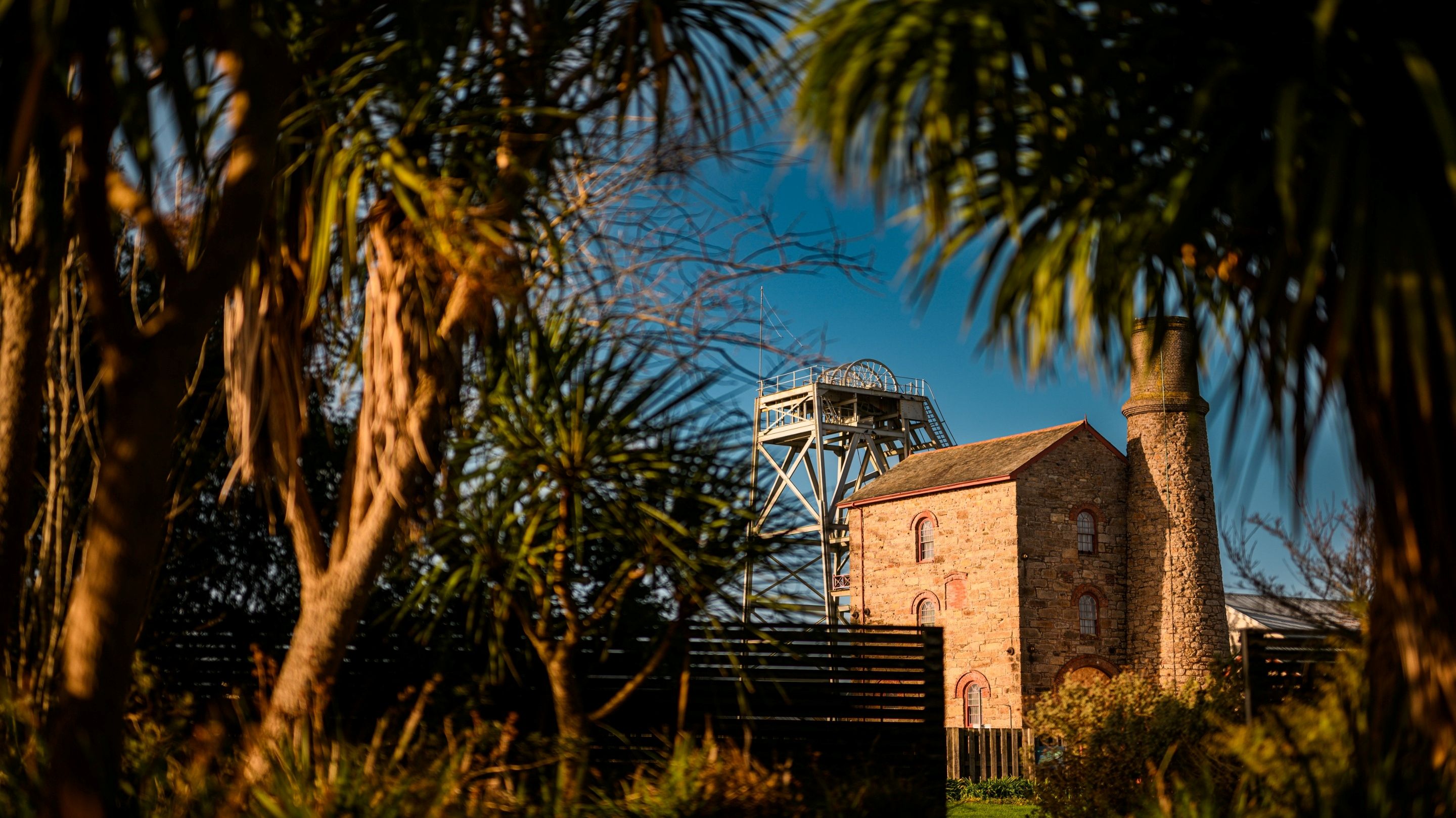 An old tall brick building with a large chimney seen through gardens with tall trees