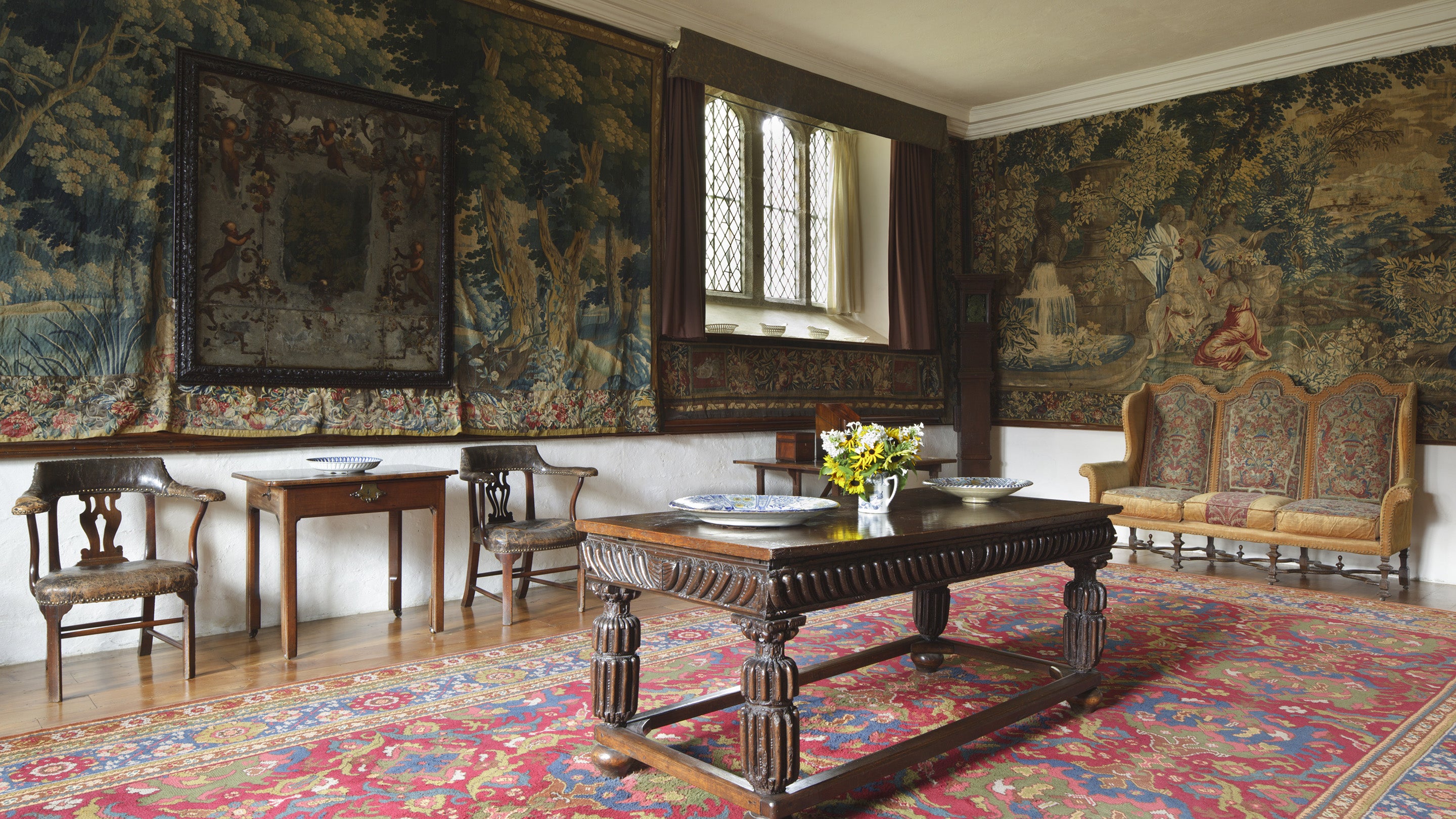 A view of a dining room, with a table in the centre and chairs pushed to the wall. The walls are adorned with a tapestry