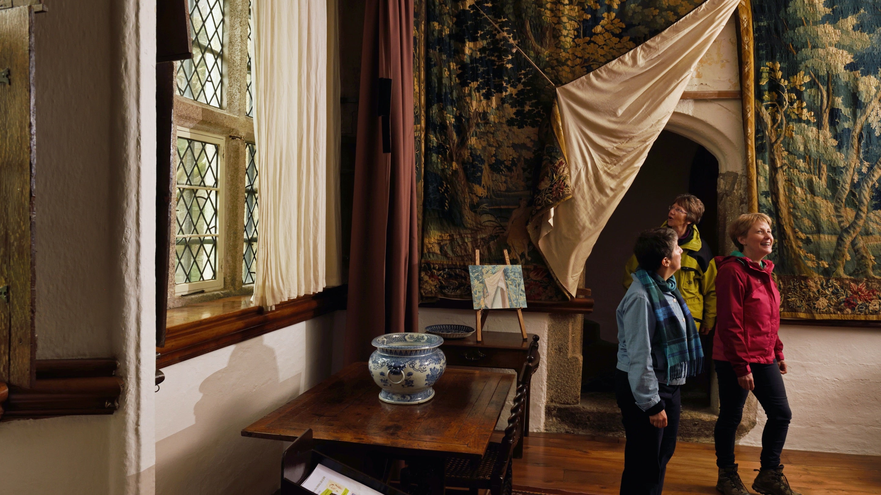 Three visitors walk through a doorway past an ornate tapestry inside the house at Cothele, Cornwall