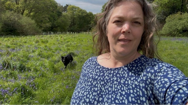 Katie, in a blue dress, walks with her black dog Lottie running behind in a grassy field