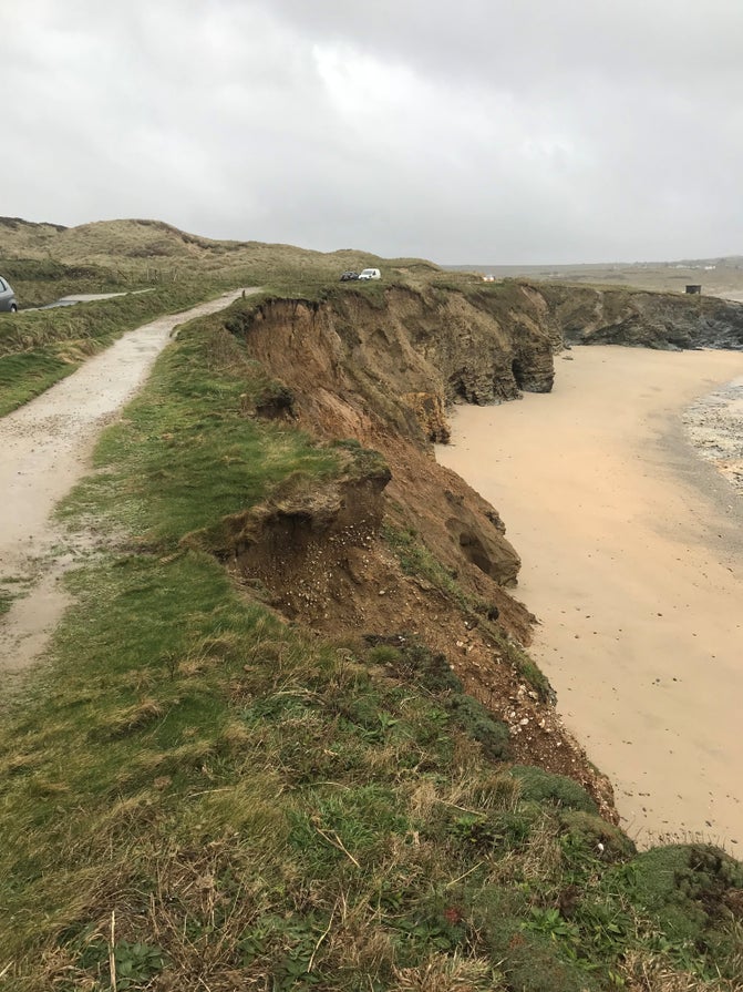 A stretch of coastline where a narrow footpath and parked vehicles sit close to the edge of a steep, eroding cliff. Fresh breaks in the exposed cliff face indicate recent erosion, highlighting the landscape’s instability. Below, a sandy beach contrasts with the fragile ground above, illustrating both the natural beauty of the coastline and the risks erosion poses to access and infrastructure. This image was taken in 2017, before the boardwalk was installed in 2018, highlighting the need for the work.