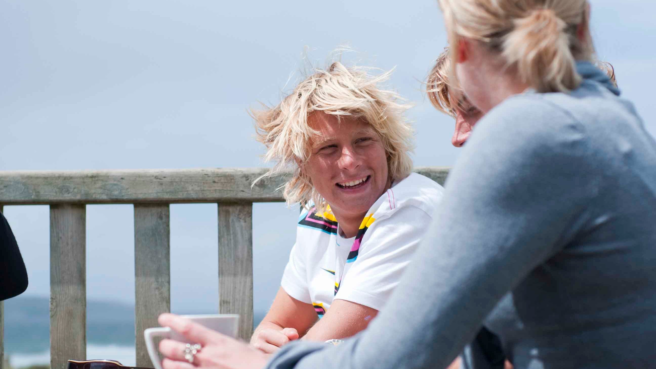 A man sitting on a bench smiling with the sea in the background and a woman with a coffee looking away from the camera