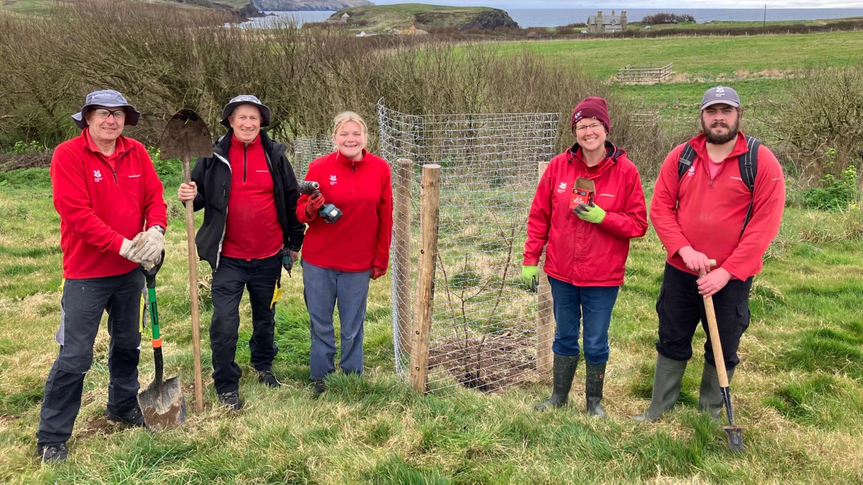 A group of five National Trust rangers planting trees smile at the camera wearing their red uniform