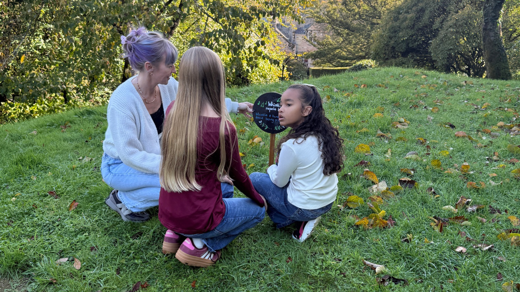 A woman and two girls crouch on the grass beside a Whistle Reeds activity sign in Lanhydrock's gardens, surrounded by trees and fallen autumn leaves.