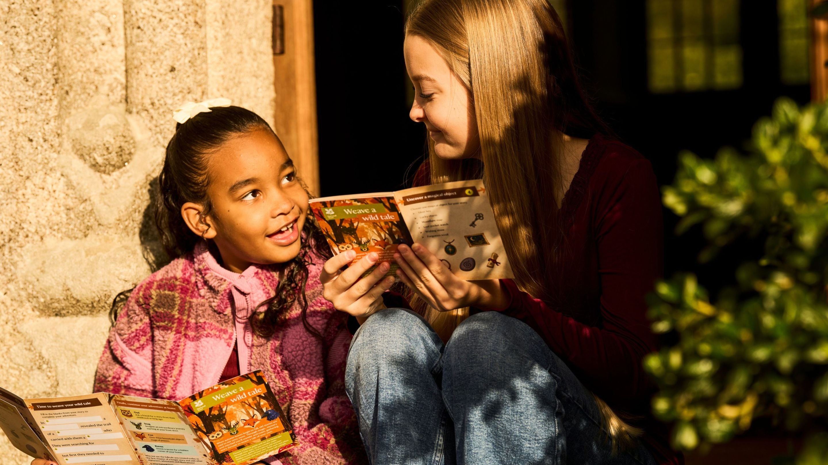 Two girls sitting together in a sunny doorway at Lanhydrock, smiling and exploring the illustrated Weave a Wild Tale autumn activity leaflets.