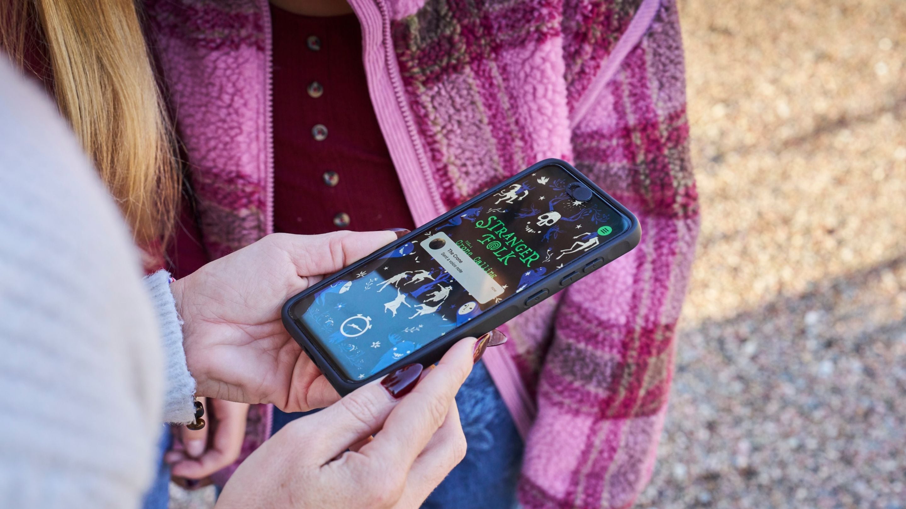 Close-up of a person holding a smartphone displaying the Stranger Folk interactive adventure, with another person wearing a pink patterned fleece standing nearby.