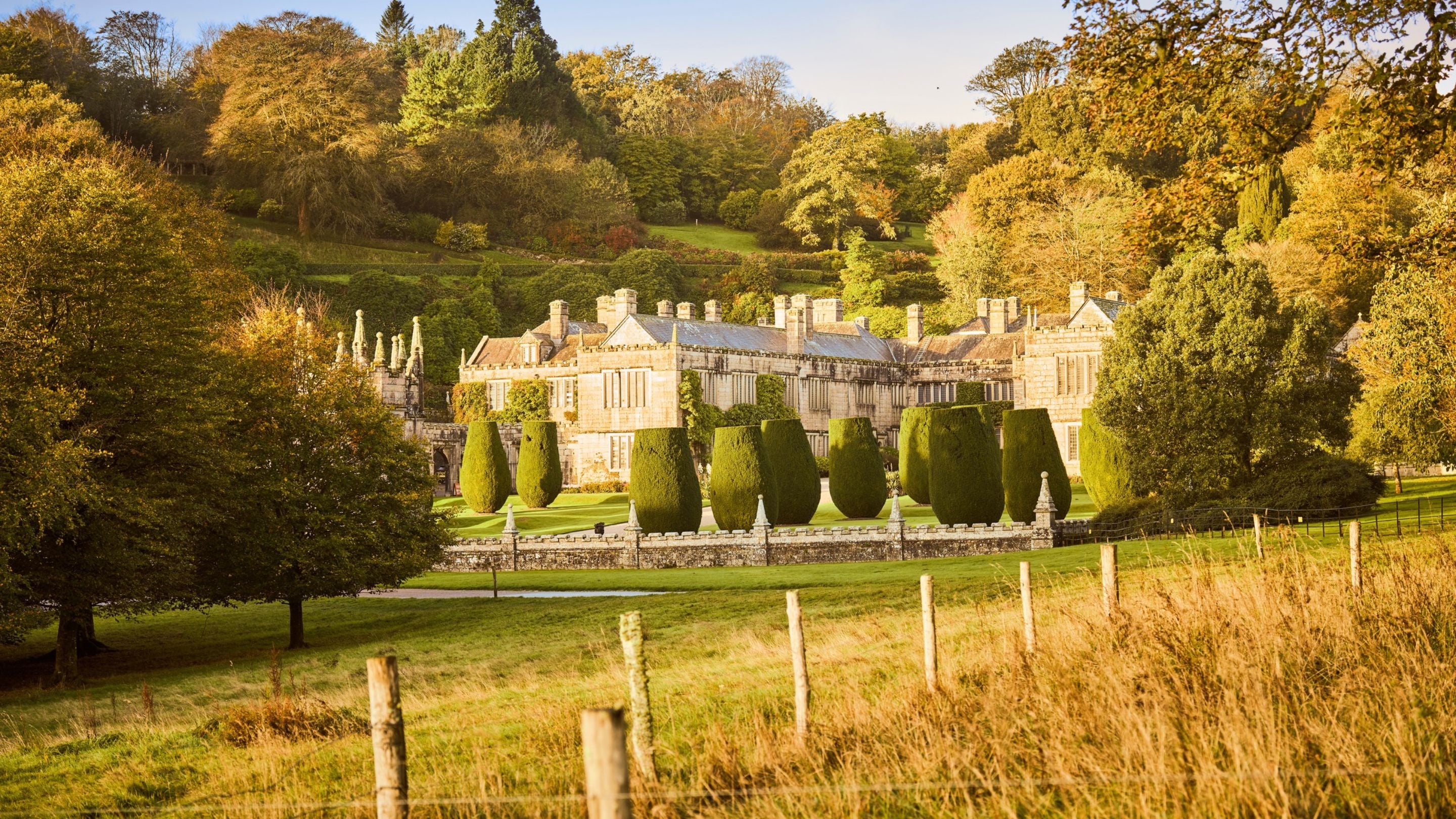 View across the parkland towards Lanhydrock house and its formal gardens, with neatly clipped yew trees, golden autumn foliage and wooded hills behind.