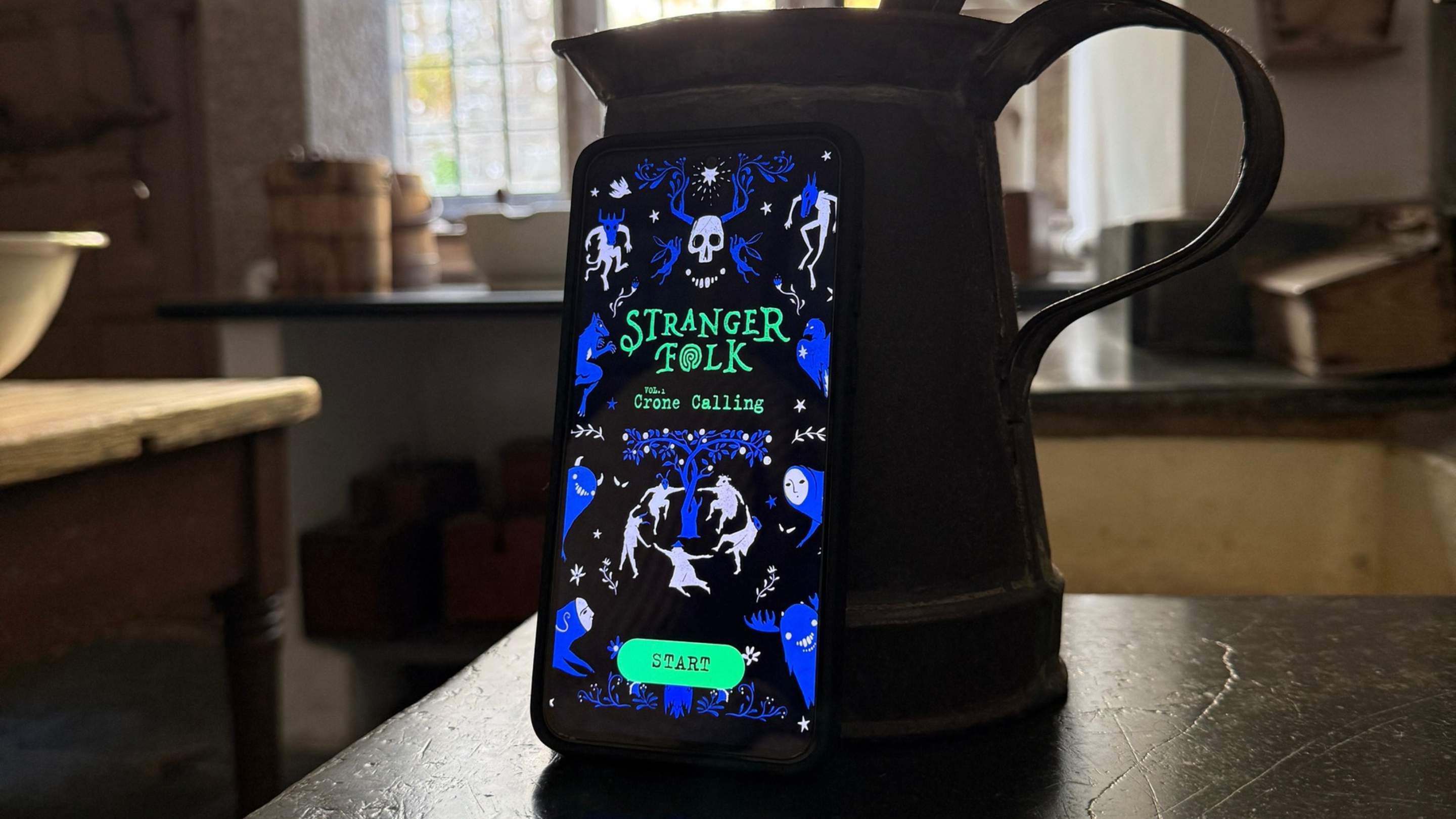 A smartphone displaying the Stranger Folk: Crone Calling app, propped against a large black jug in Lanhydrock’s historic kitchen.
