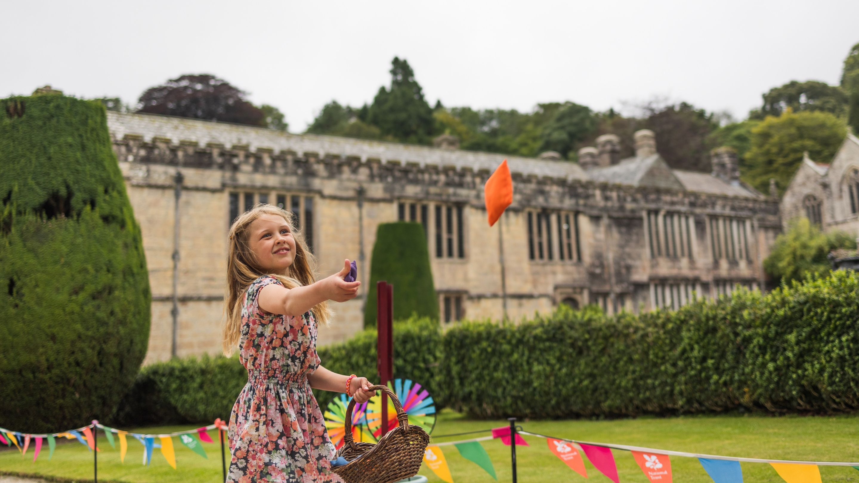 A girl in a pink flowery dress throwing a bean bag into the air in front o Lanhydrock house